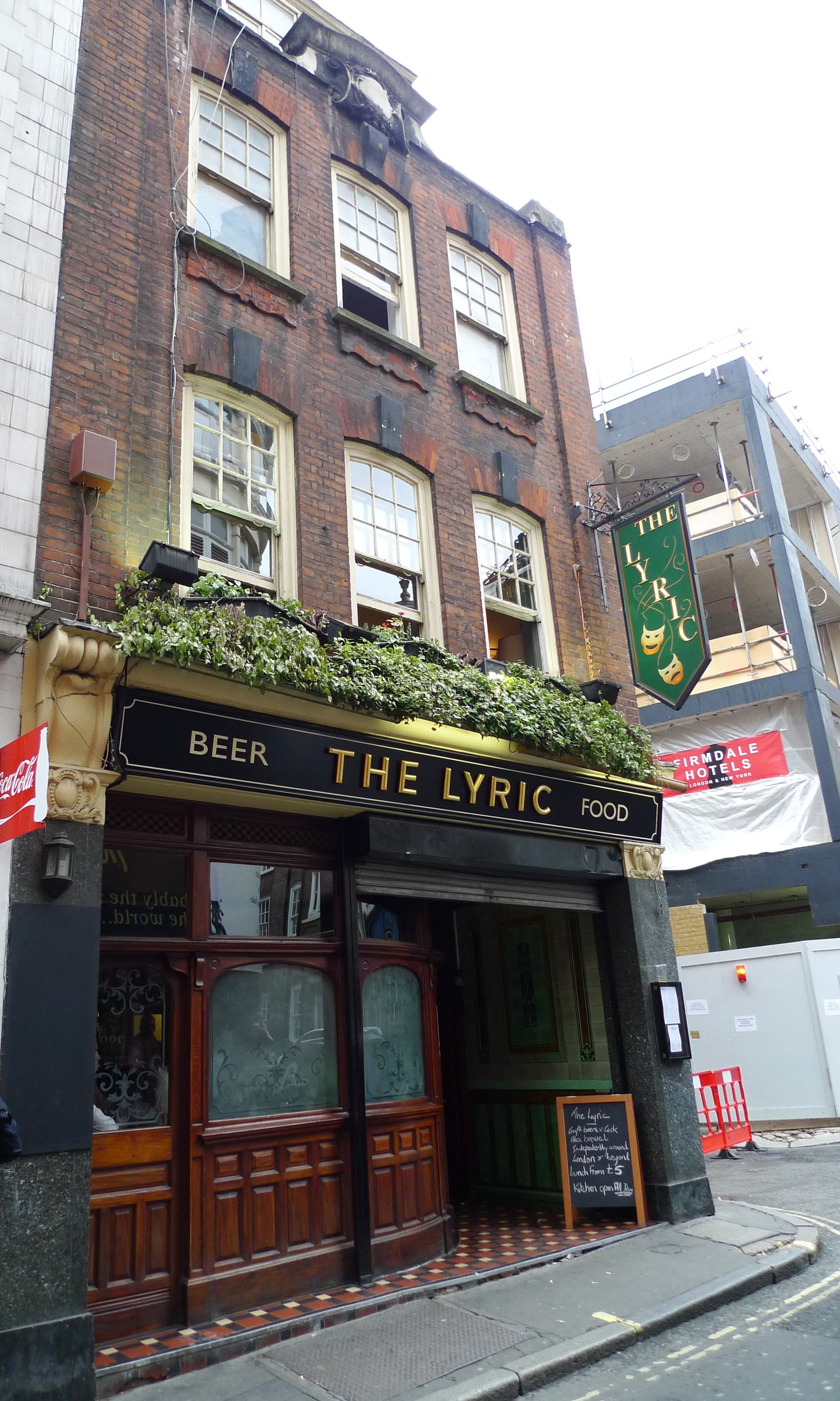 Brick frontage of The Lyric pub in Soho with black fascia, ivy, hanging sign and tiled entrance