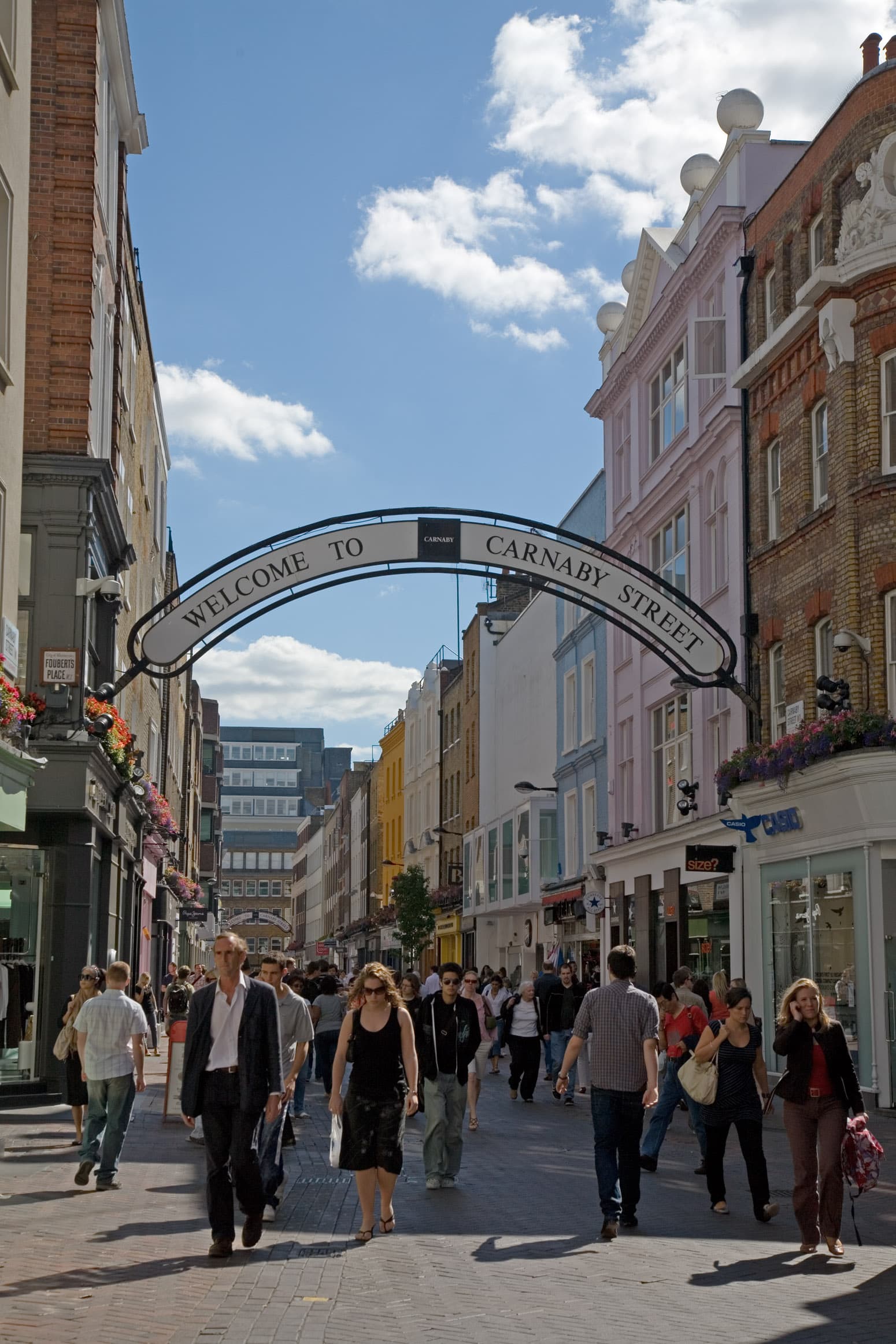 Pedestrians walk beneath the Welcome to Carnaby Street arch, with colourful shopfronts and buildings lining the pedestrianised Soho street