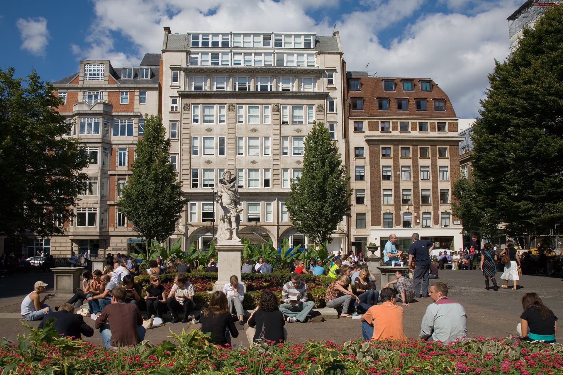 Office workers eating lunch on the grass and paving around the statue in Golden Square, Soho, on a sunny day
