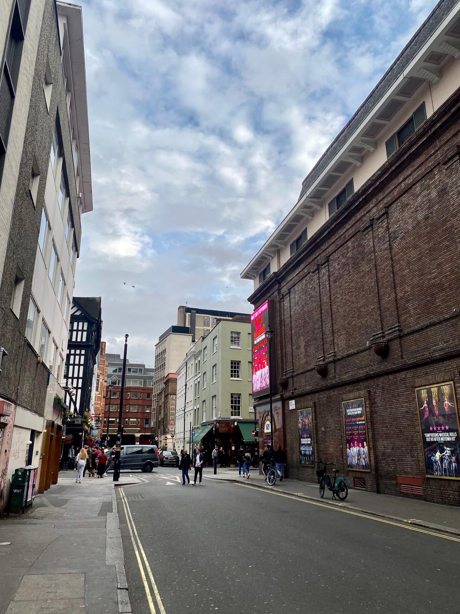 Soho street corner with brick theatre wall, pub frontage, pedestrians and a cloudy sky near Old Compton Street