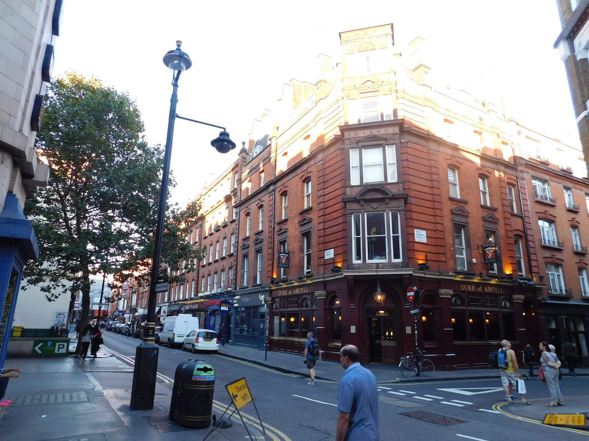 Red-brick corner pub and shopfronts on Brewer Street, Soho, with pedestrians, parked vehicles and a lamppost in evening light