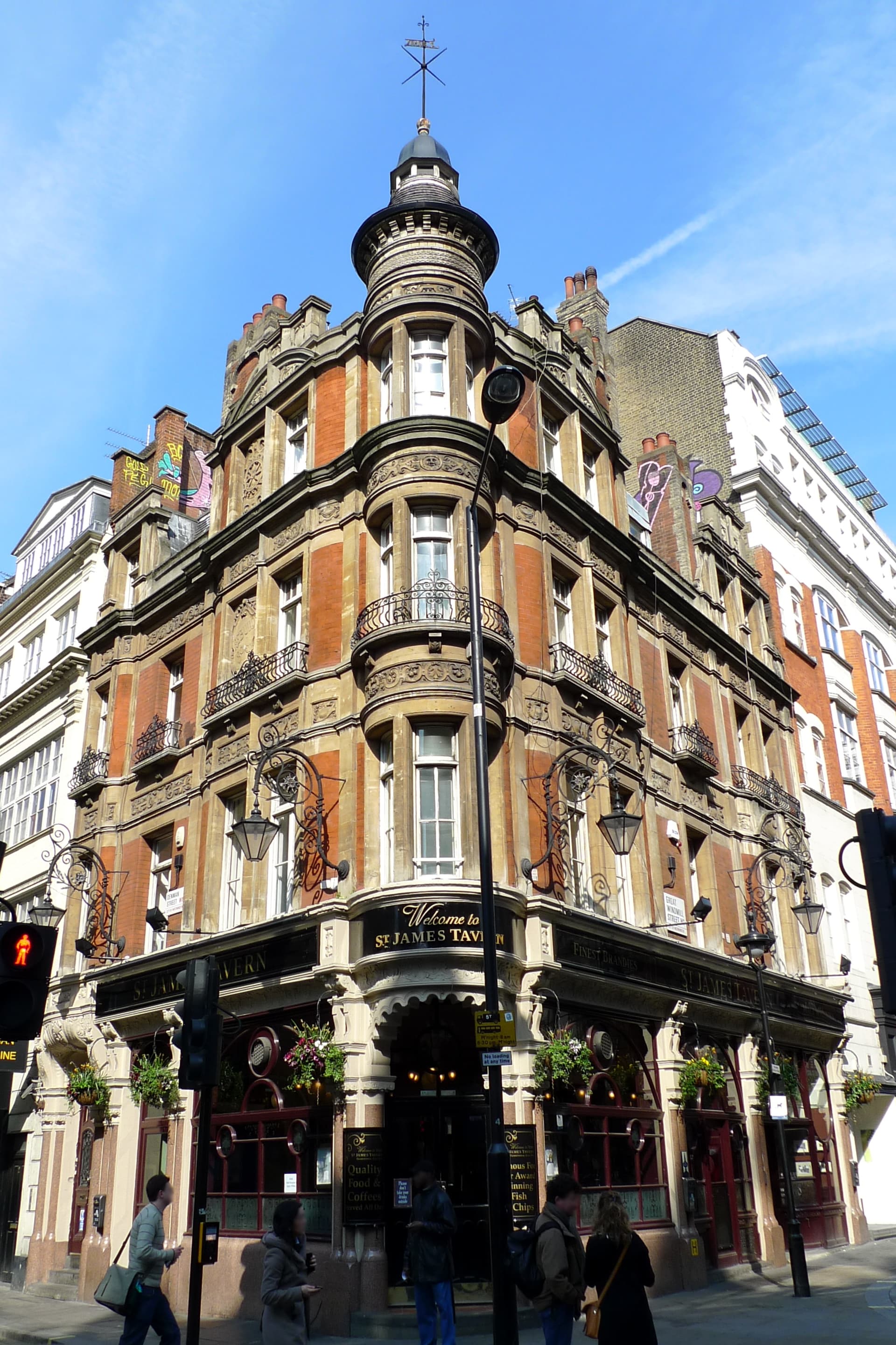 Ornate Victorian red-brick and stone corner pub with turret and wrought-iron lamps on a sunny Soho street corner