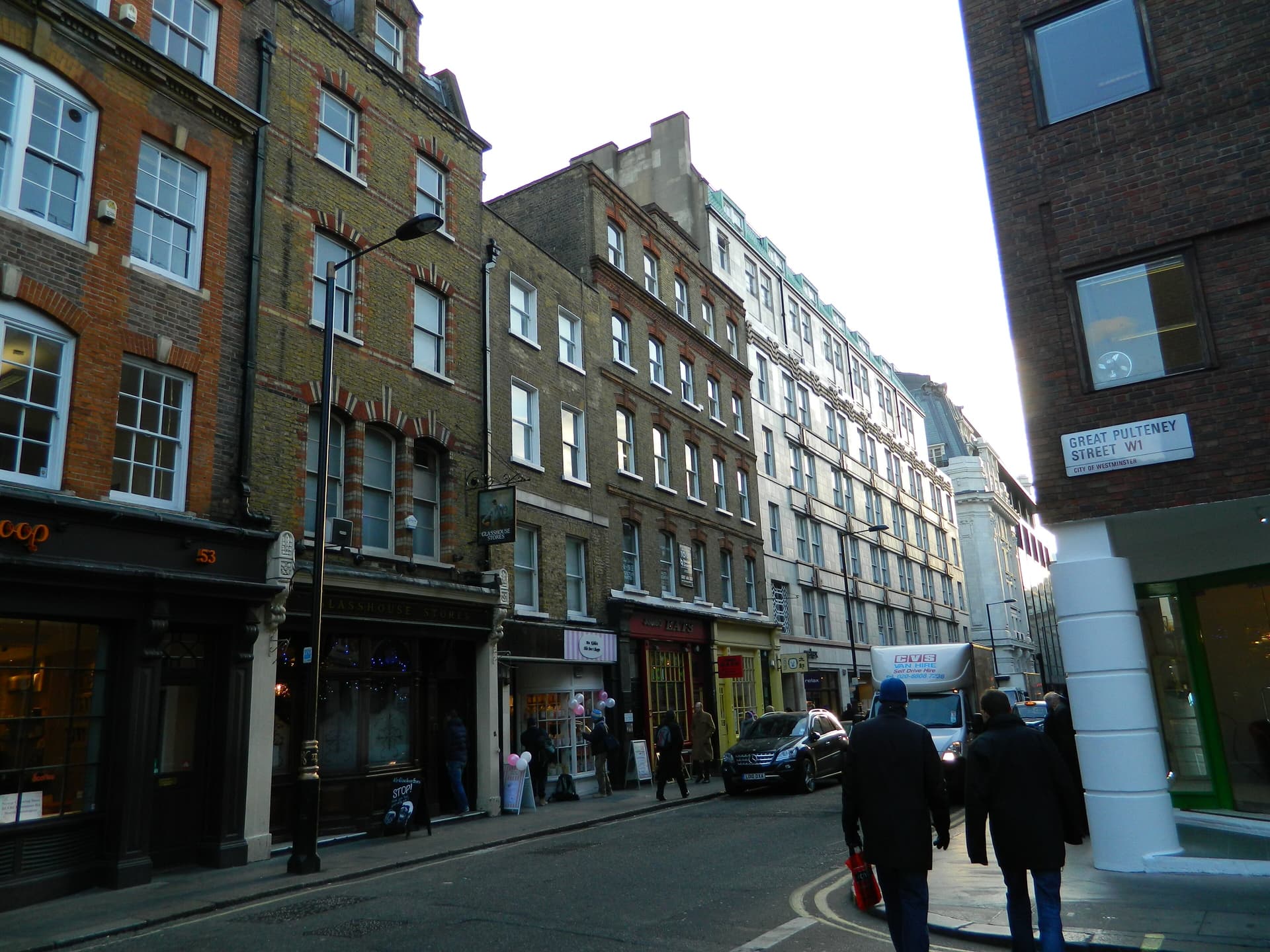 Victorian brick buildings and small shopfronts along a Soho street, with pedestrians and a van near Great Pulteney Street
