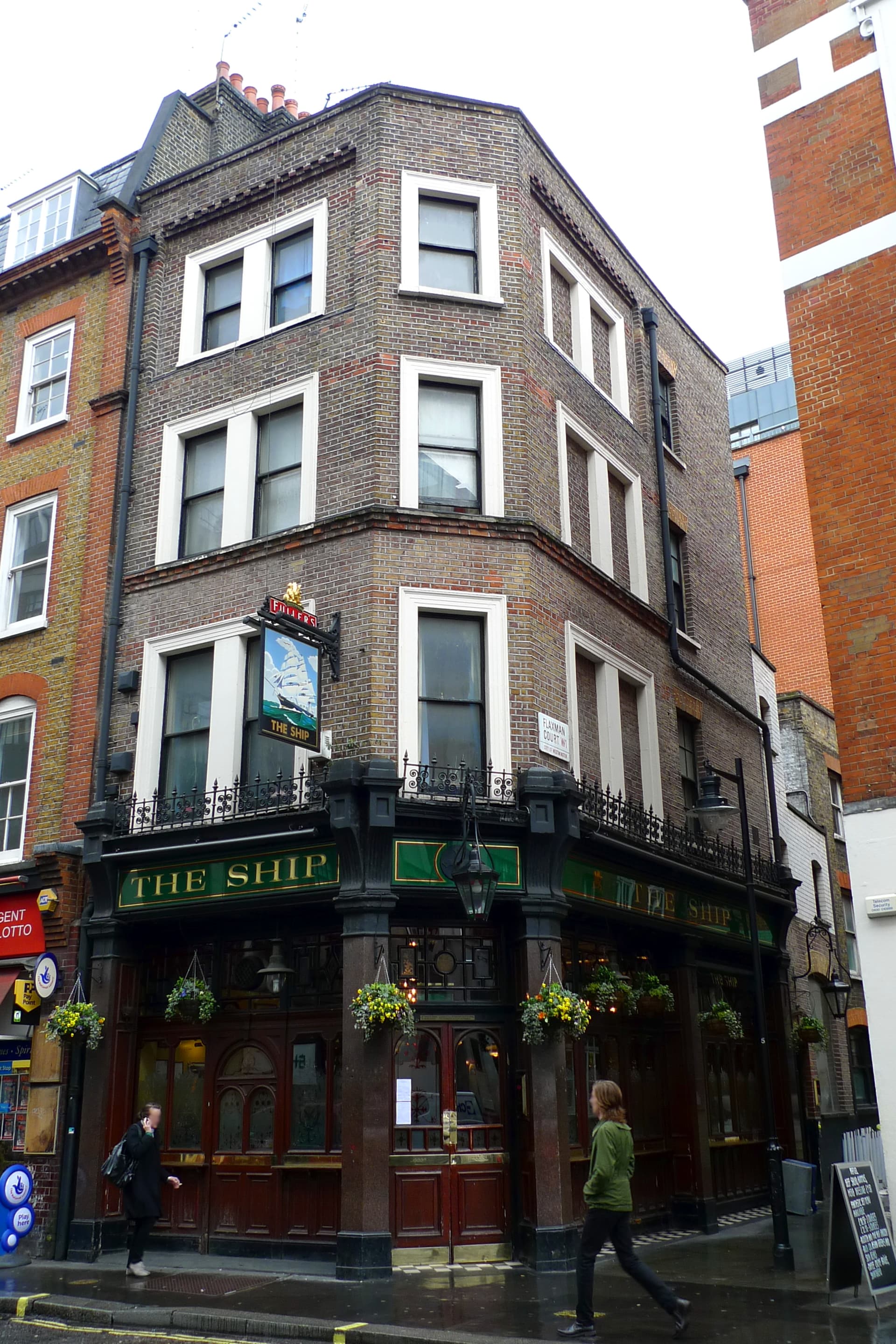 Corner pub The Ship with dark green fascia, hanging flower baskets and brick upper floors on a wet Soho street