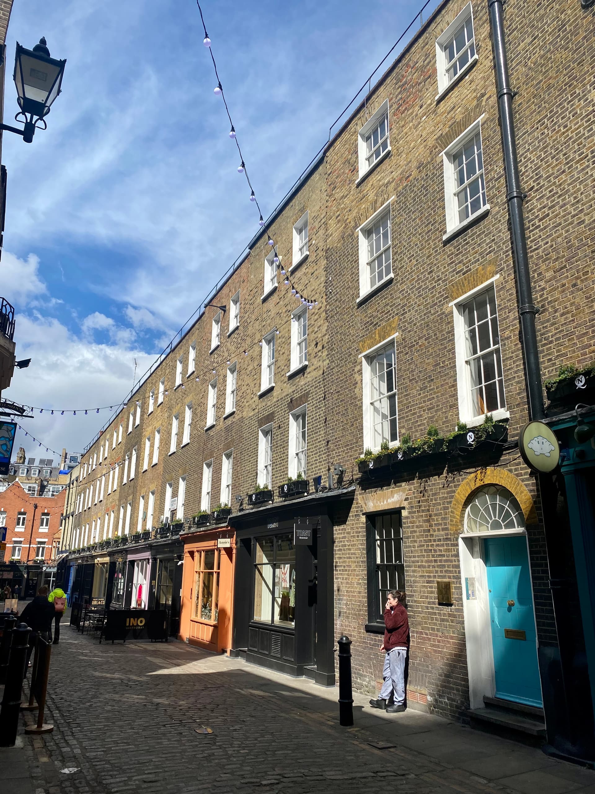 Sunlit Georgian brick terrace with colourful shopfronts, cobbles and festoon lights along a narrow Soho street in London