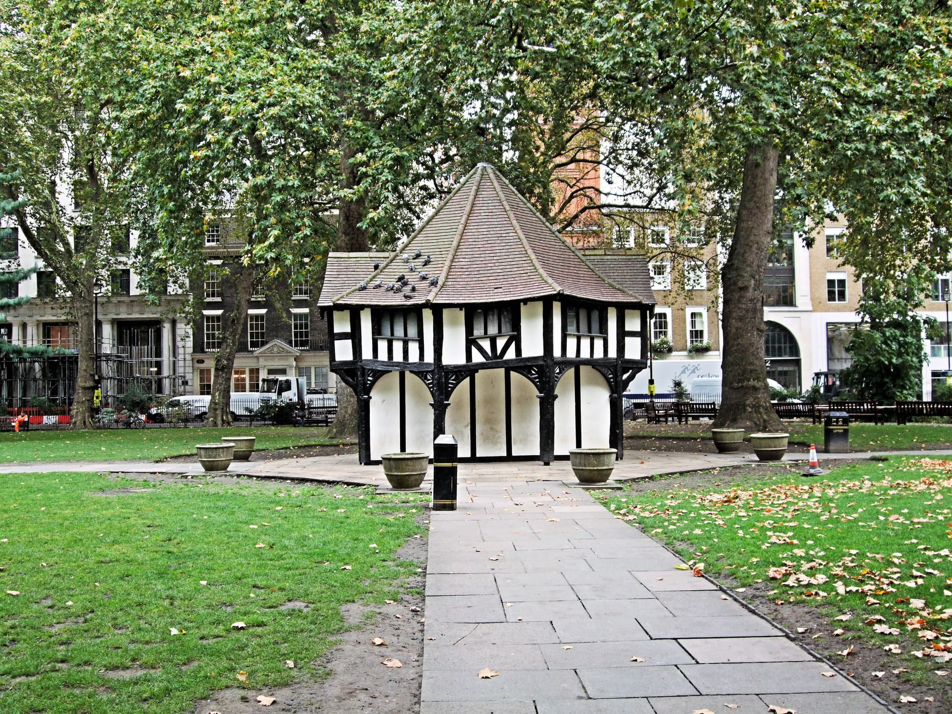 Mock-Tudor timber-framed garden hut with pigeons on its roof, surrounded by plane trees and Georgian buildings in a London square