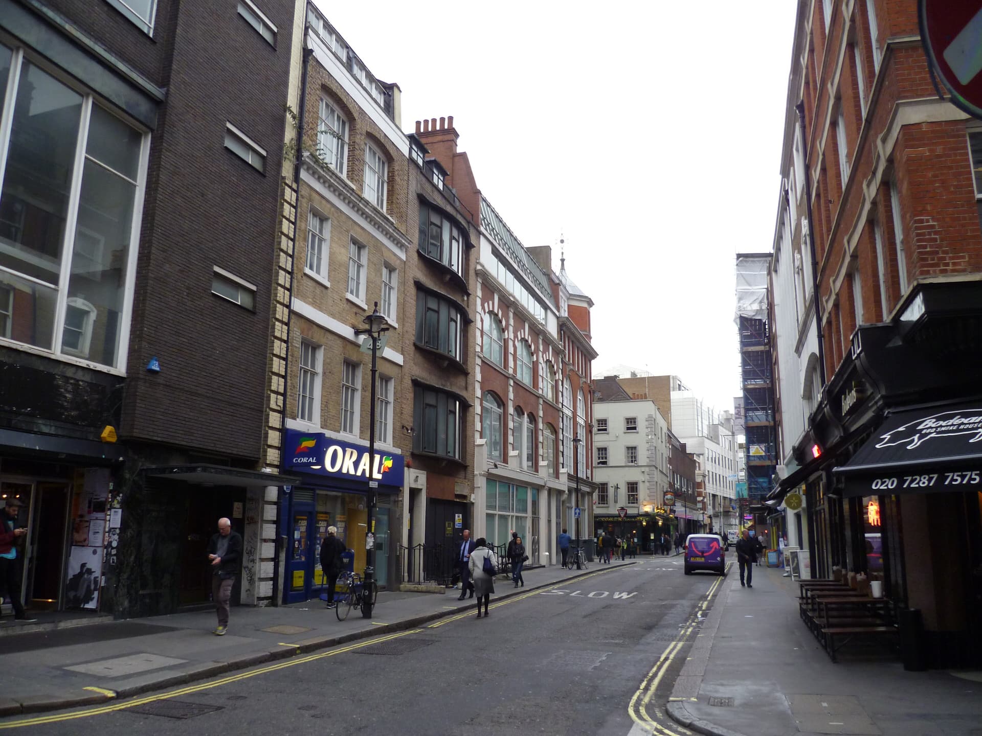 Soho street of mixed brick buildings with a bookmaker's, a barbecue restaurant awning and pedestrians under an overcast sky