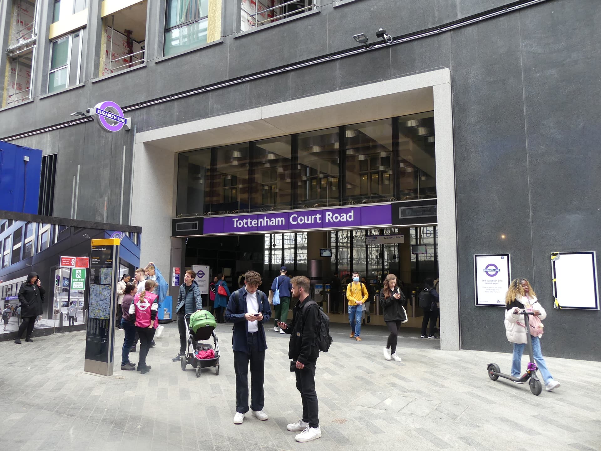 Elizabeth line entrance at Tottenham Court Road station, with pedestrians, a pushchair and a scooter rider on the paved forecourt