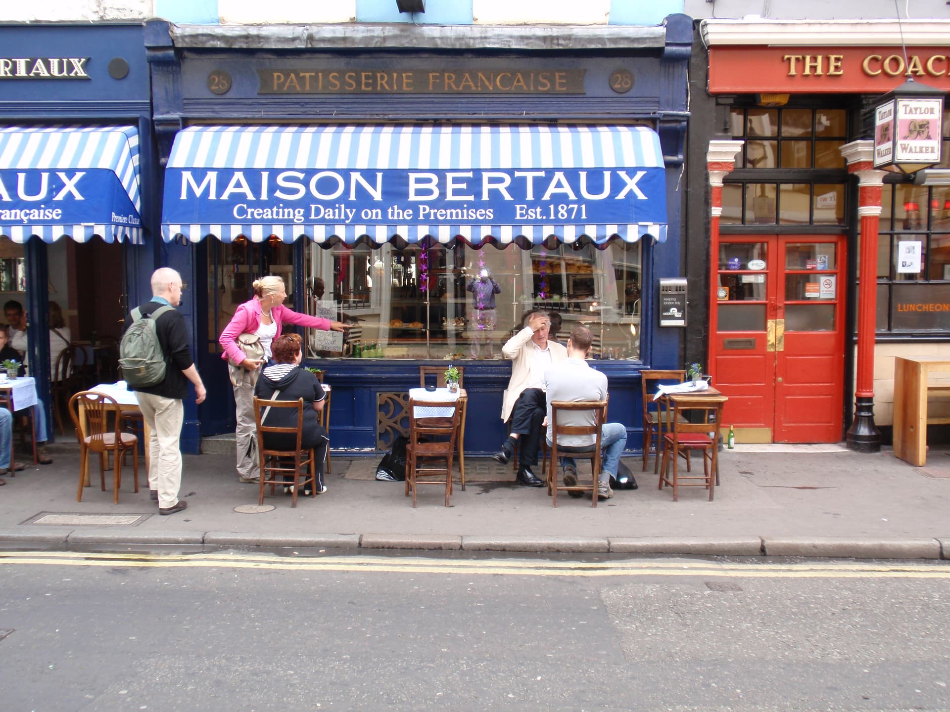 Blue-fronted Maison Bertaux patisserie with striped awning and pavement tables beside a red-fronted pub on a Soho street