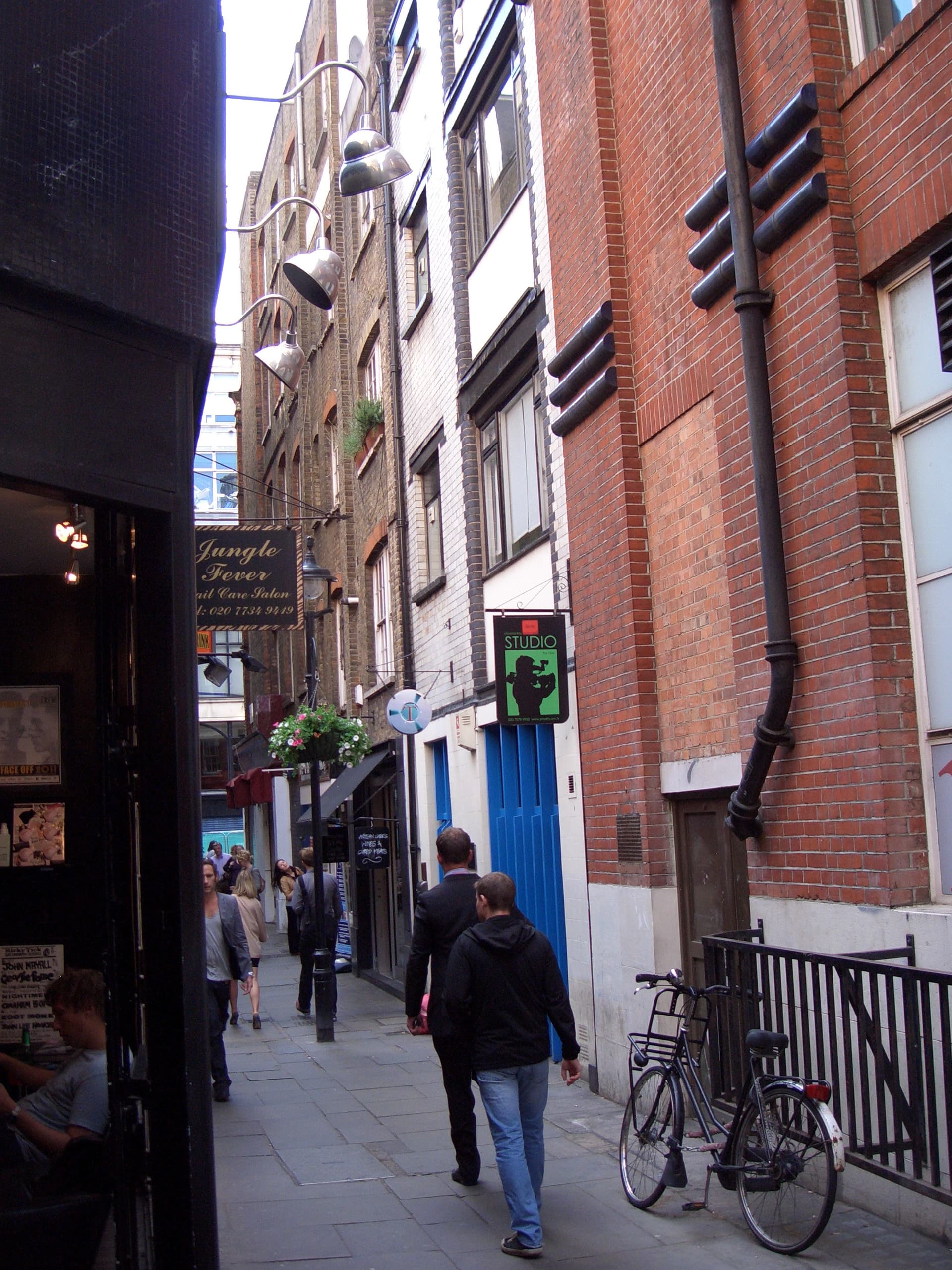 Narrow Soho alley with brick buildings, hanging signs, a parked bicycle and pedestrians walking along the paving