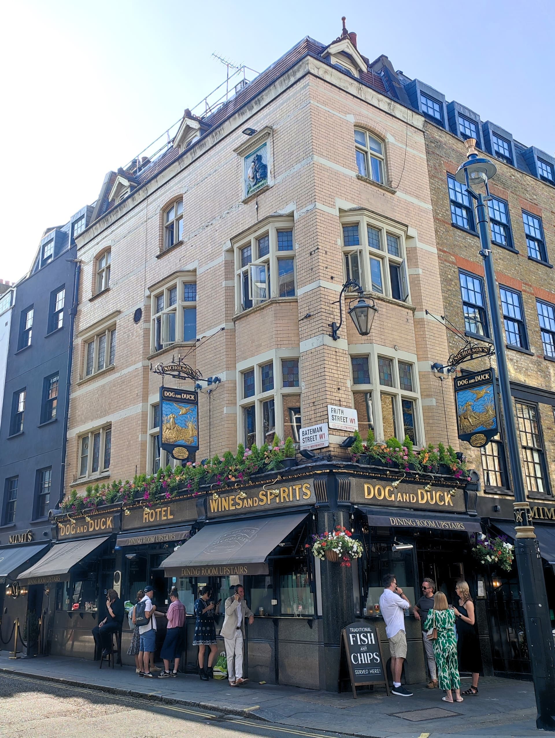 Corner Victorian brick pub with gold lettering, hanging signs and flower boxes, drinkers standing outside on a sunny Soho street