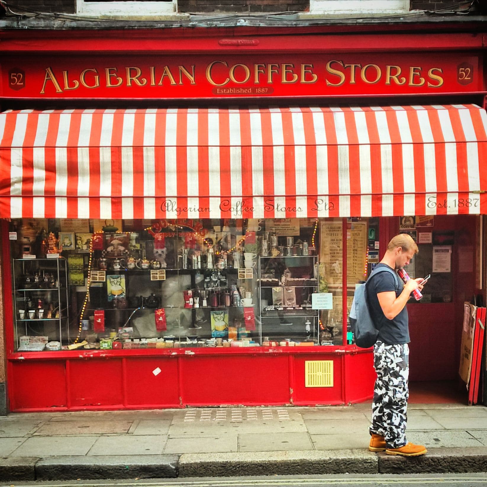 Red-fronted Algerian Coffee Stores with striped awning and crowded window display, a man in camouflage trousers walking past