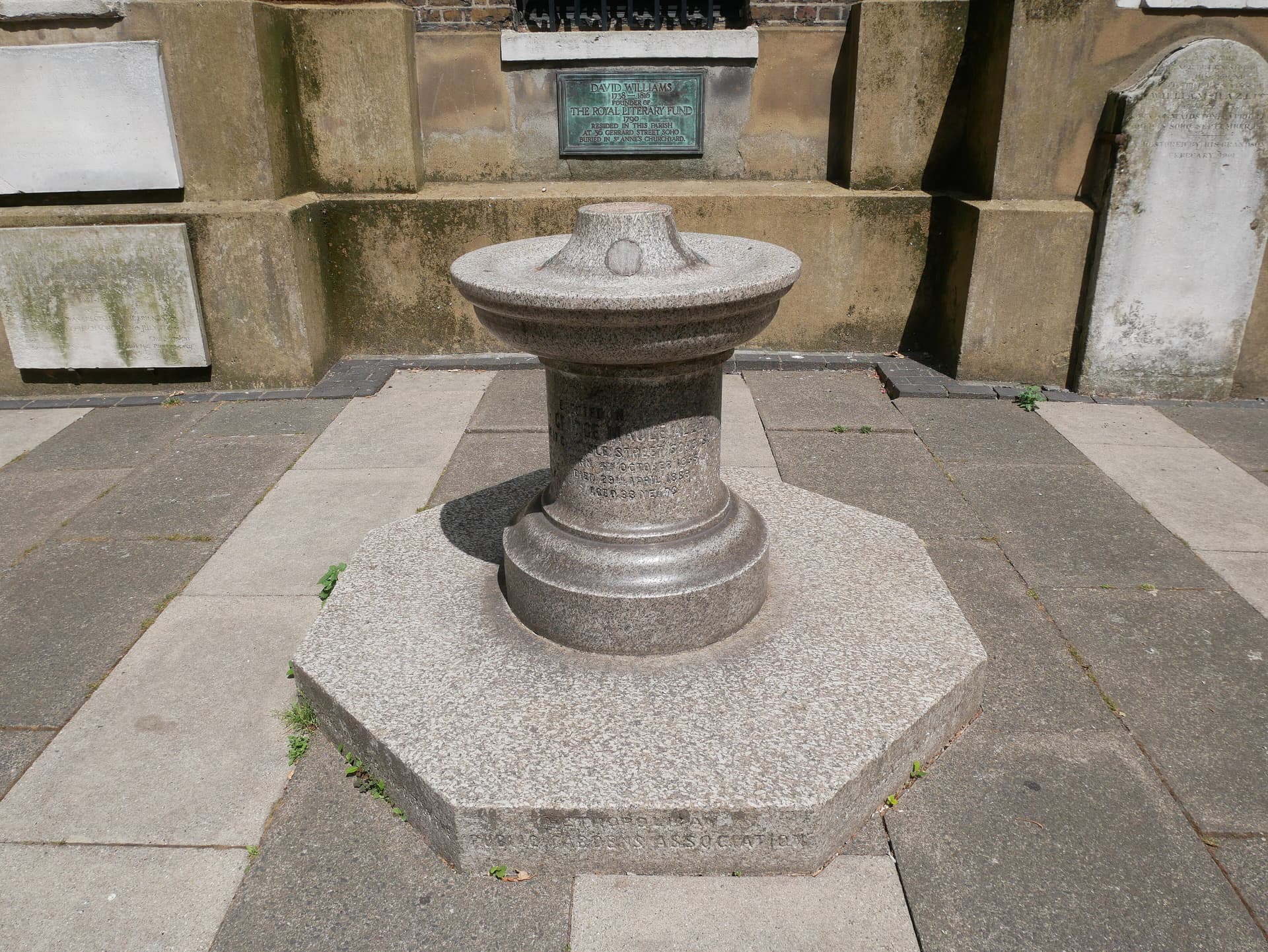 Granite drinking fountain on an octagonal plinth in sunlit St Anne's churchyard, Soho, beneath a green plaque