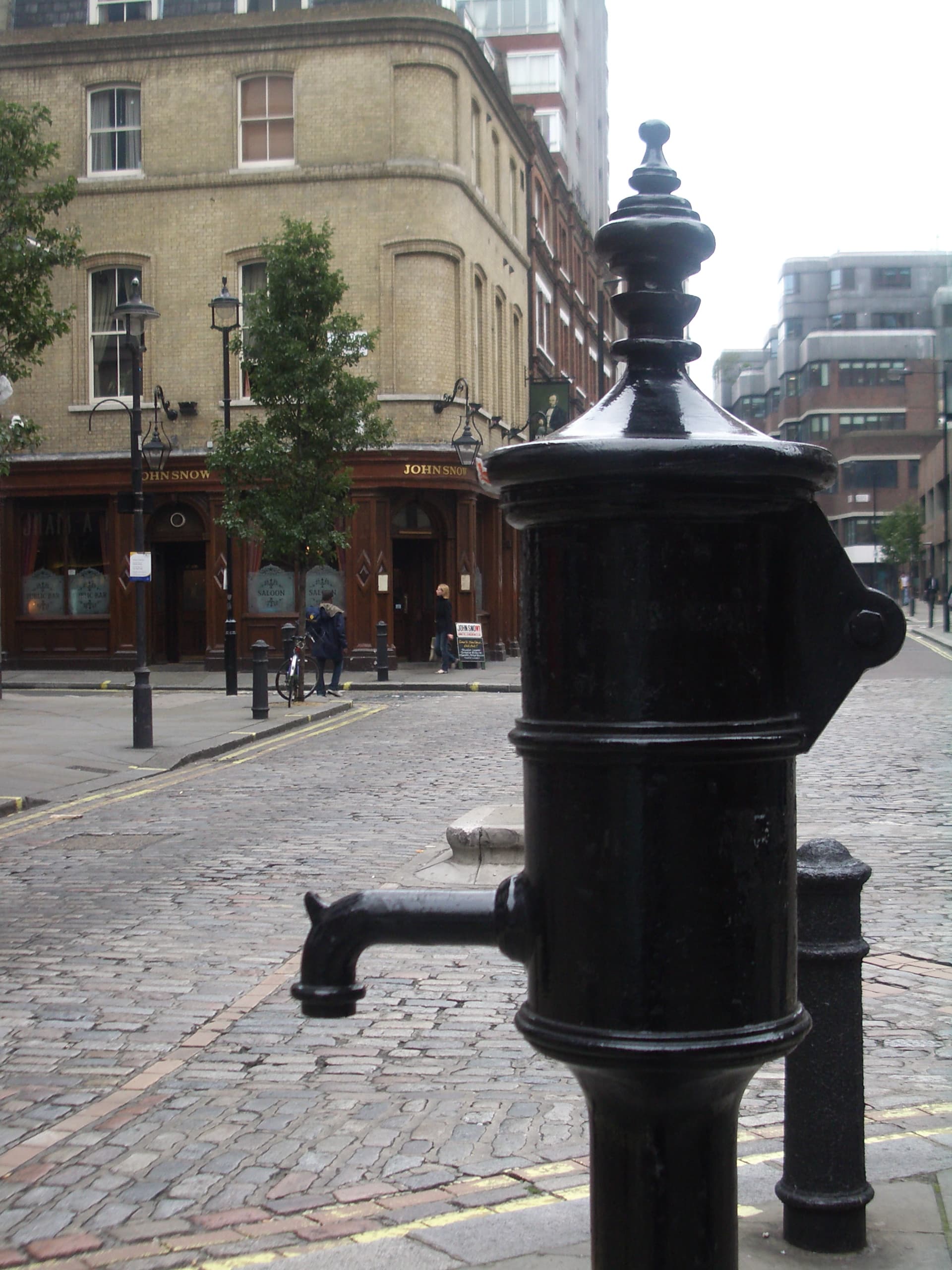 Black cast-iron replica water pump on a cobbled Soho corner, with the John Snow pub behind