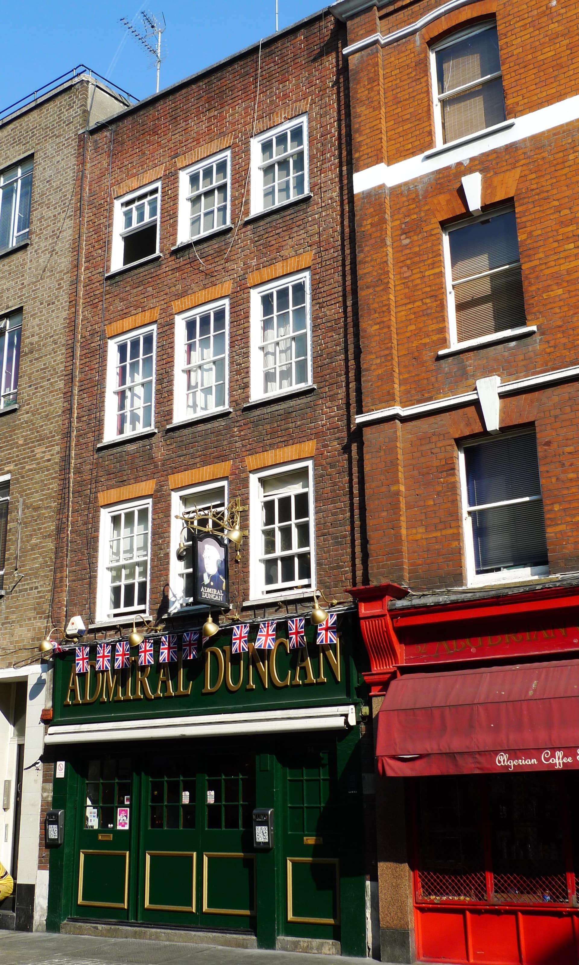 Green-fronted Admiral Duncan pub with Union Jack bunting beneath a red brick Soho townhouse, beside a red Algerian Coffee Stores awning