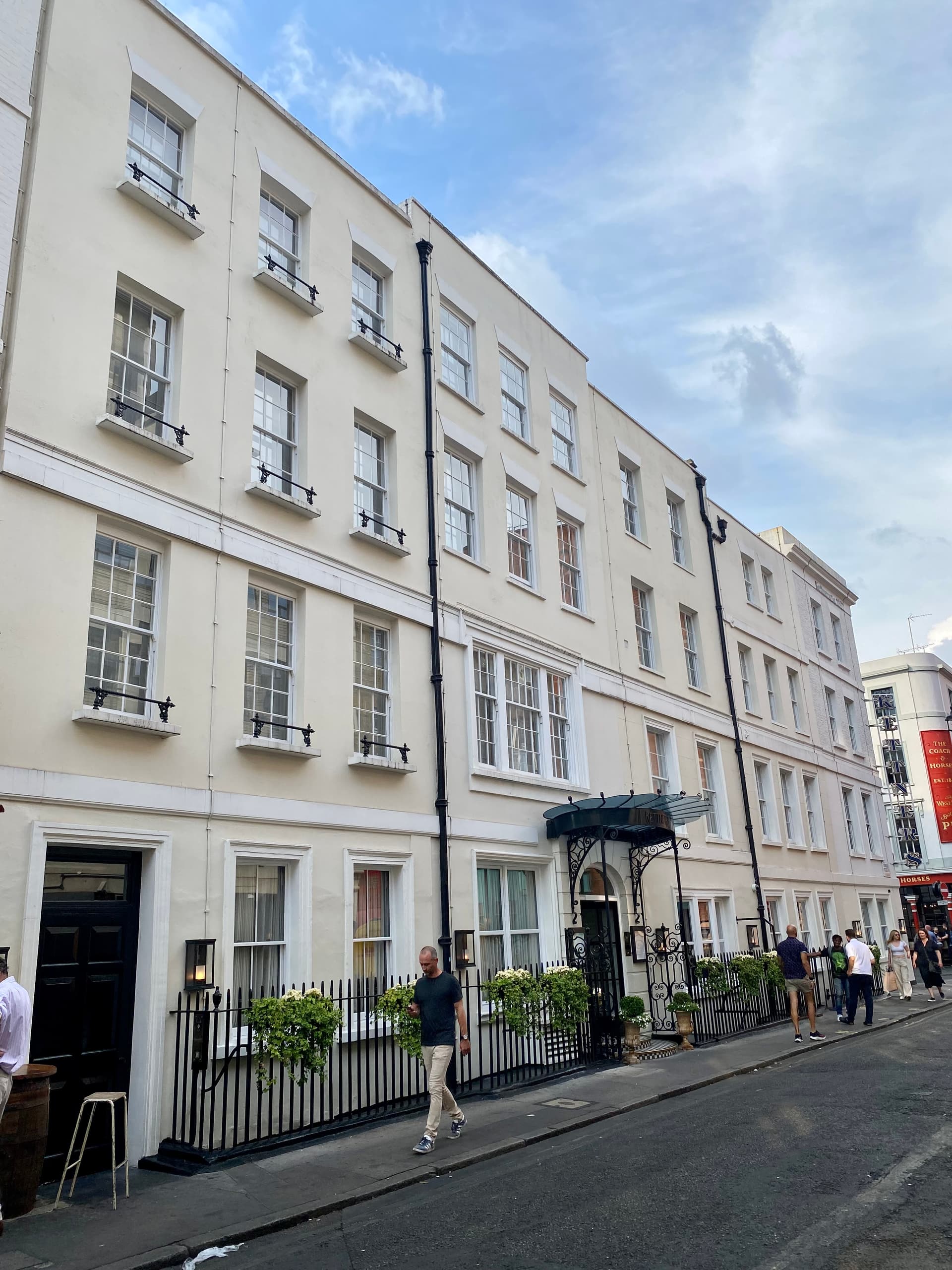 Cream stucco Georgian terrace with black railings, a glass canopy entrance and pedestrians on a Soho street
