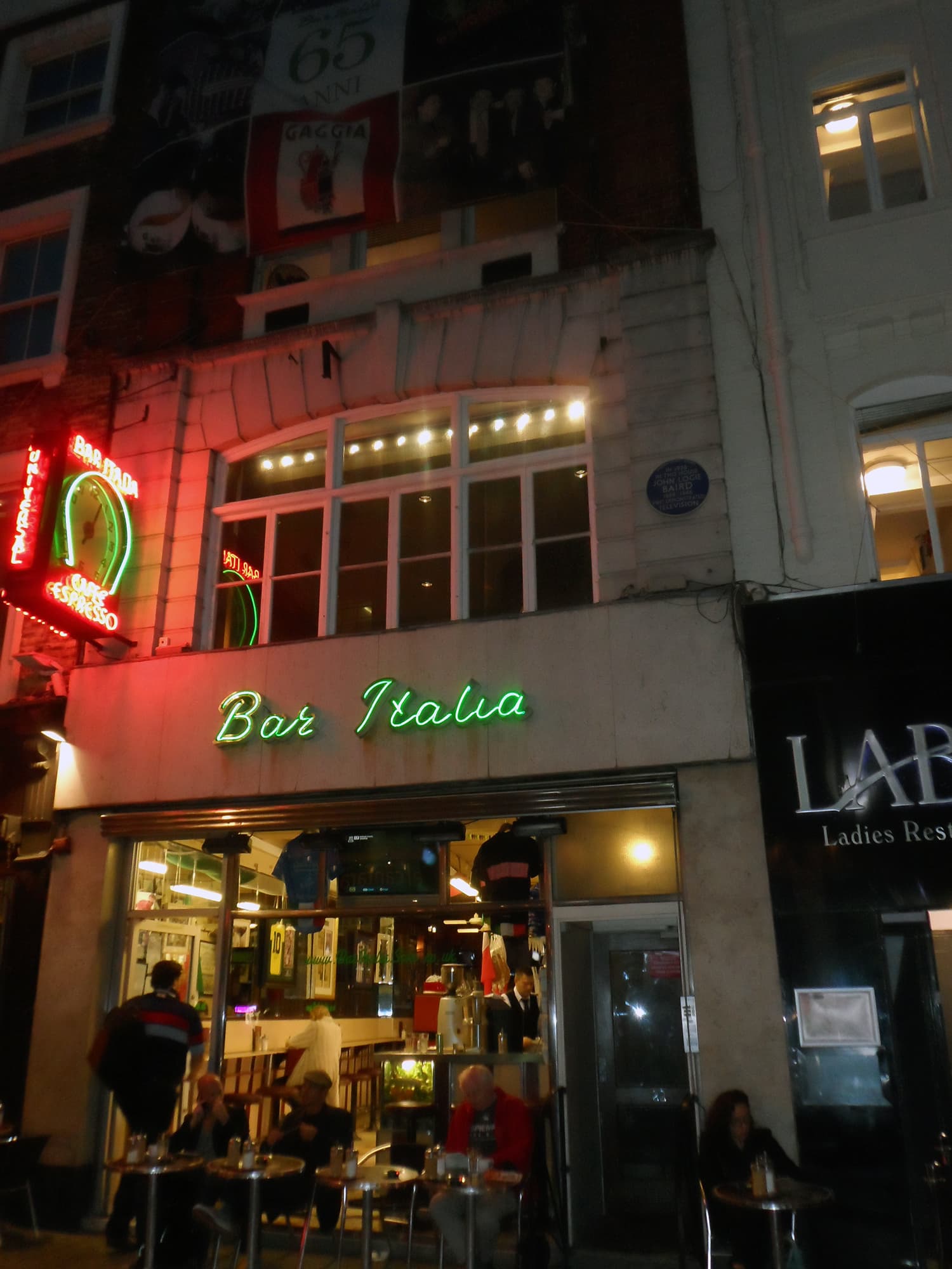 Neon-lit Bar Italia frontage on Frith Street at night, with customers at pavement tables and a blue plaque above
