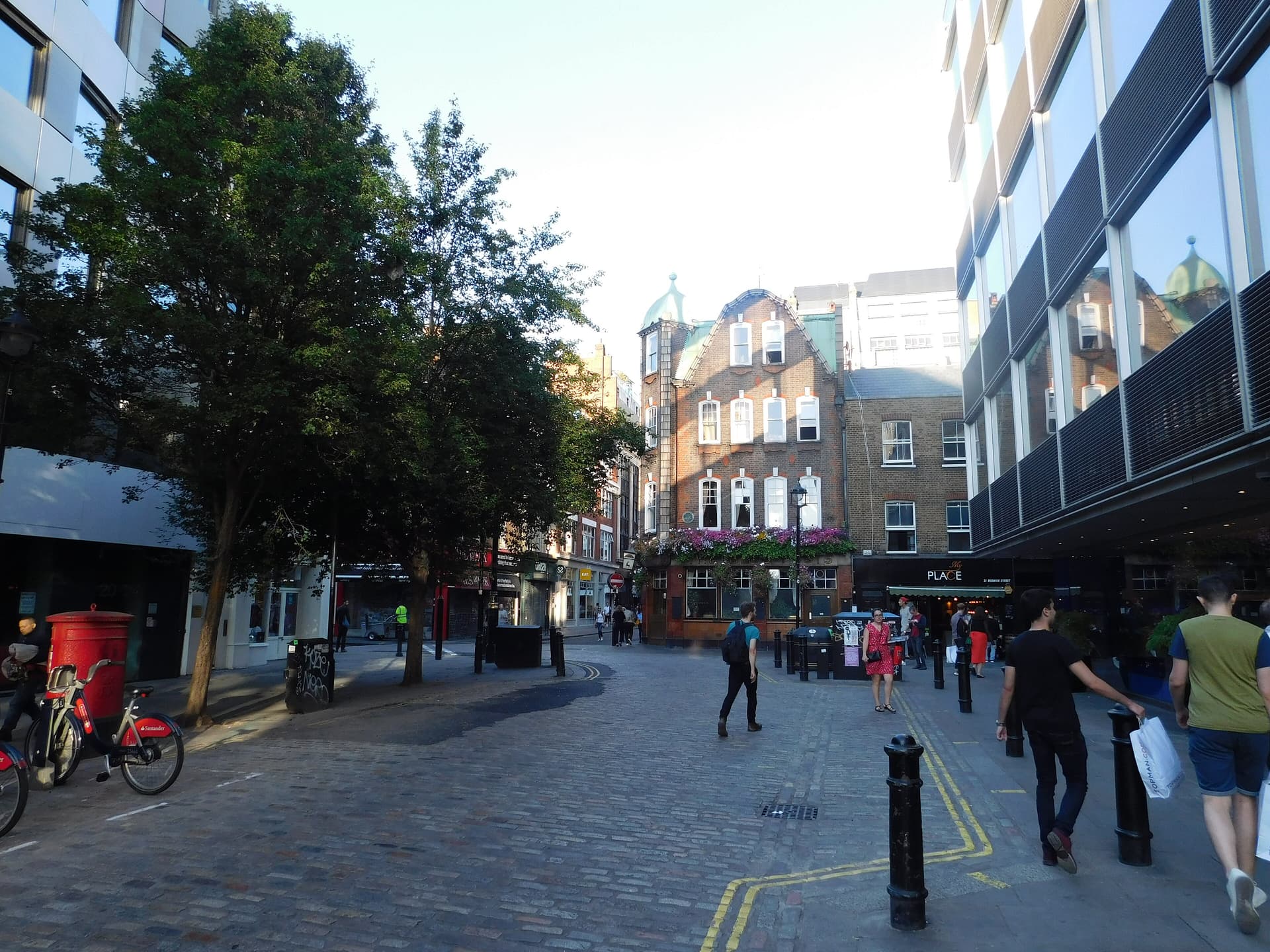 Cobbled Soho street corner with a flower-covered pub, red postbox, Santander bikes and pedestrians in evening light