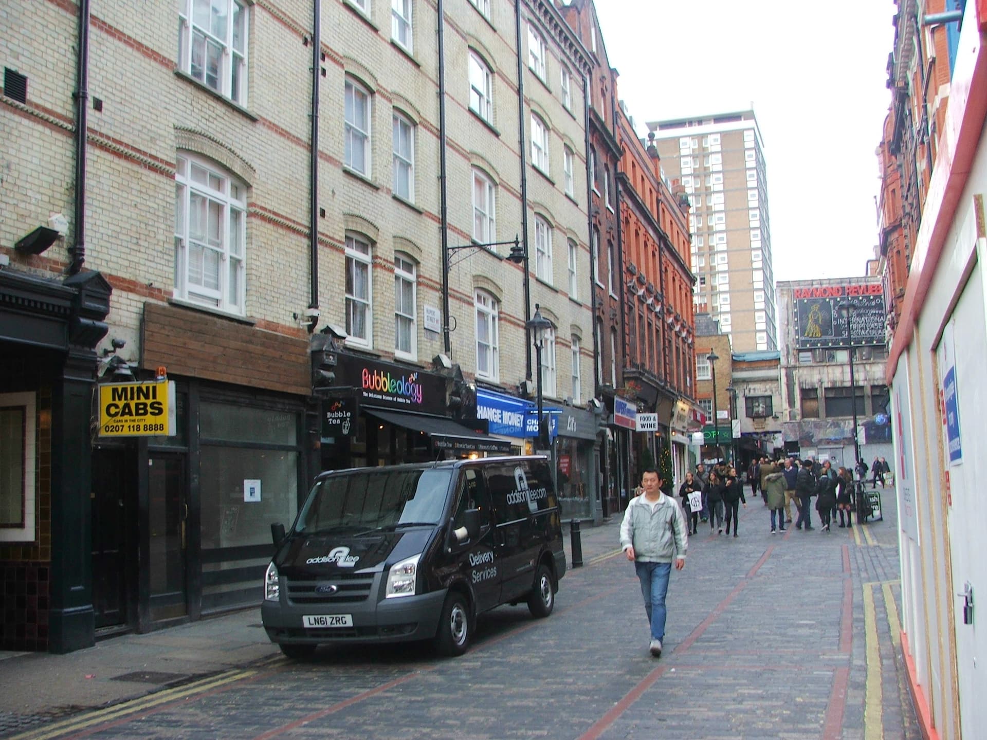 Pedestrians walk along a cobbled Soho street lined with shops, past a parked black van and the Raymond Revuebar sign