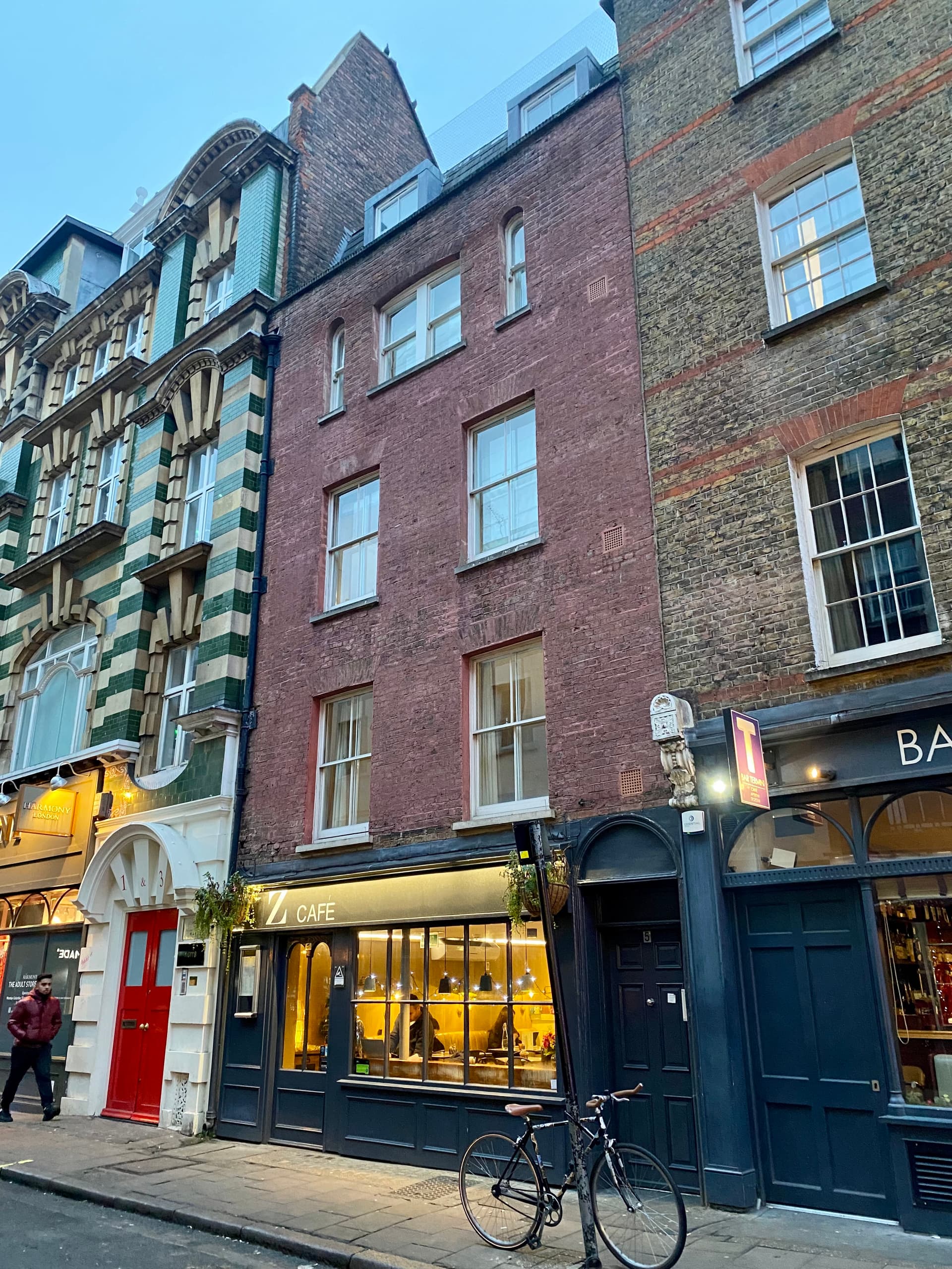 Narrow red-brick Georgian townhouse above a lit café, beside a green and cream tiled building on a Soho street