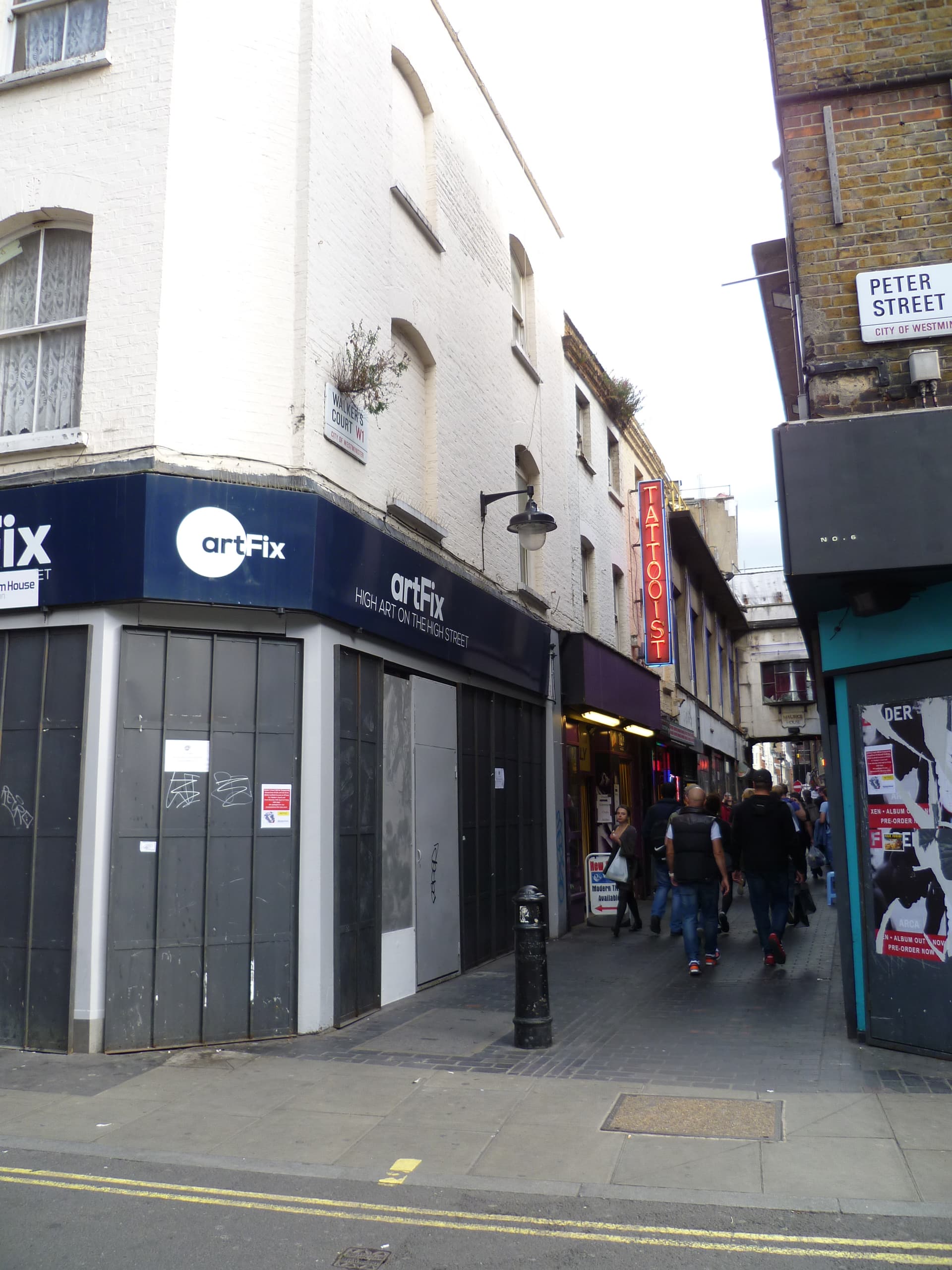 Pedestrians walk along narrow Walker's Court in Soho, past a shuttered corner shop and a red neon tattooist sign