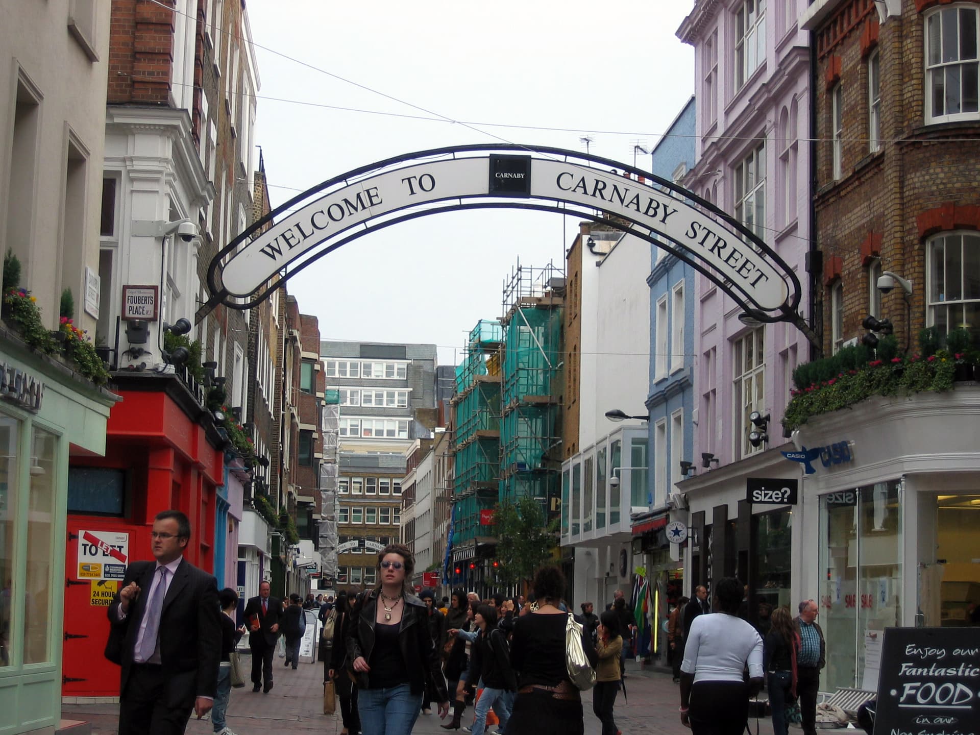 Pedestrians beneath the Welcome to Carnaby Street arch, with colourful shopfronts and scaffolding along the busy street