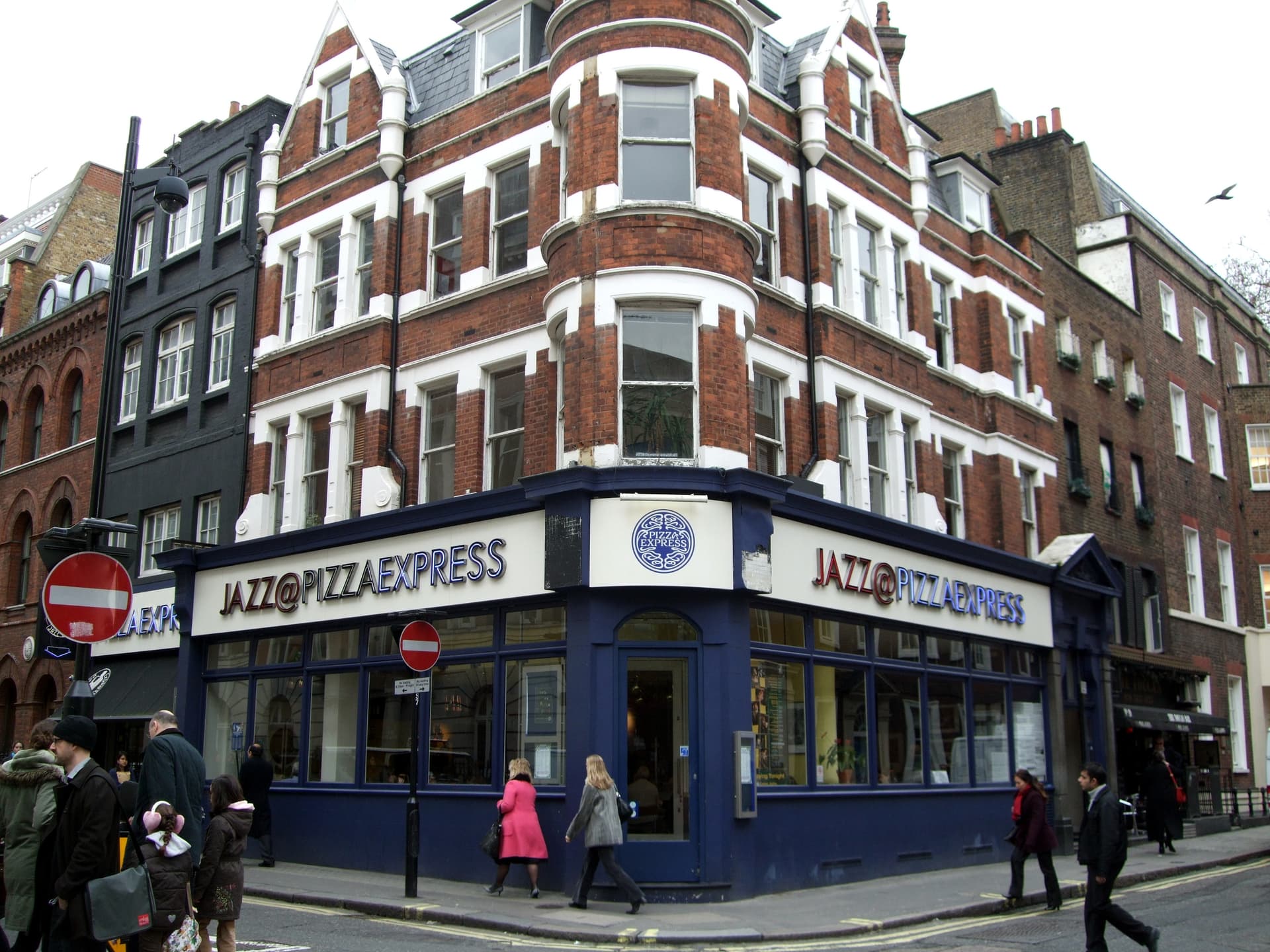 Corner red-brick Victorian building with navy shopfront signed Jazz@PizzaExpress on a Soho street, pedestrians passing