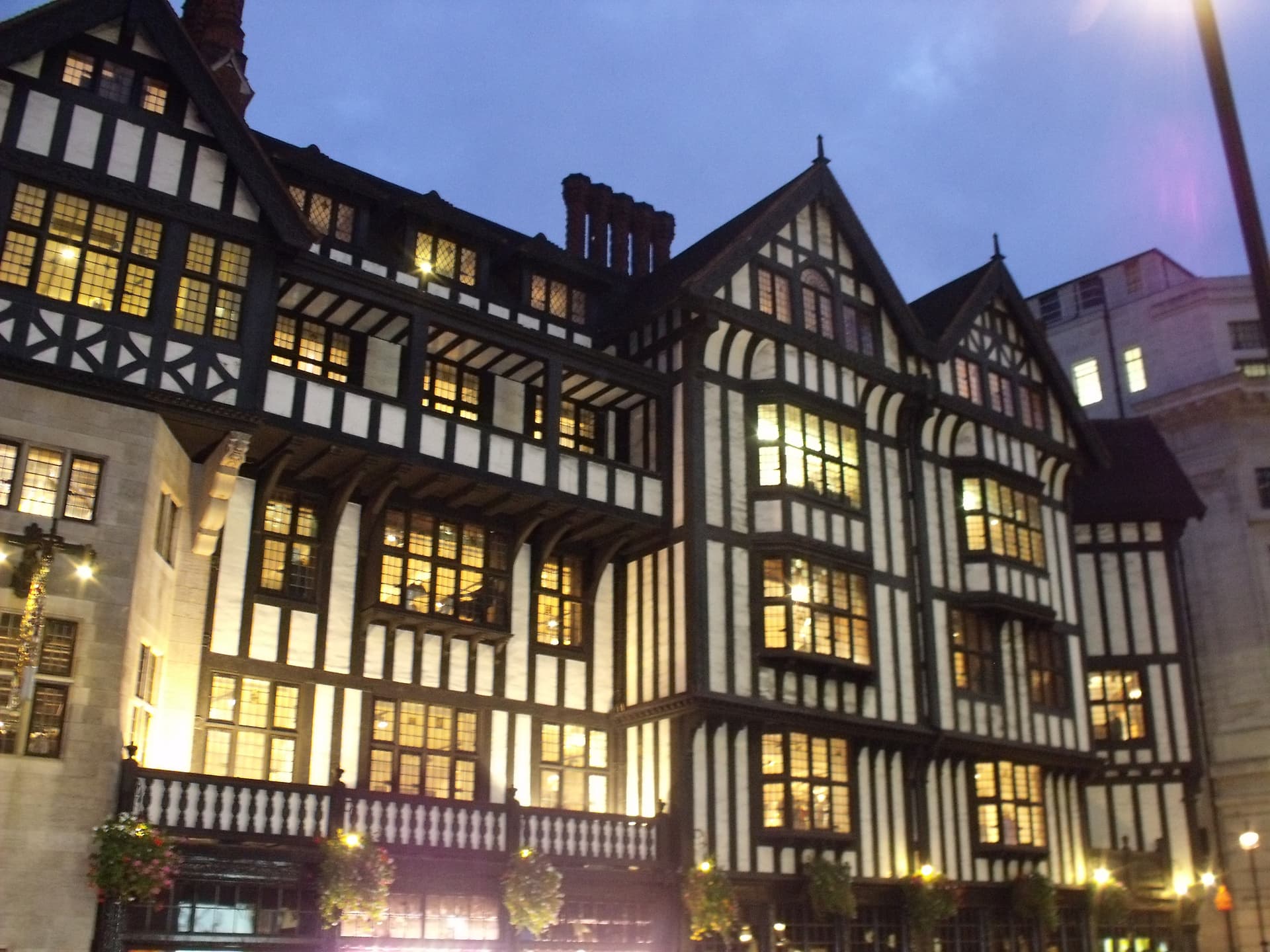 Black-and-white Tudor timber-framed building with lit leaded windows and hanging flower baskets at dusk