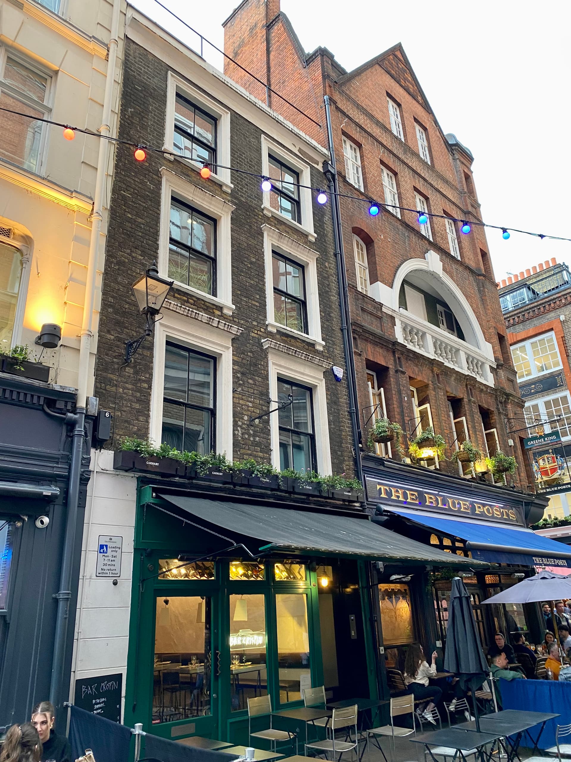 Brick townhouses above Bar Crispin and The Blue Posts pub on a Soho street strung with coloured festoon lights