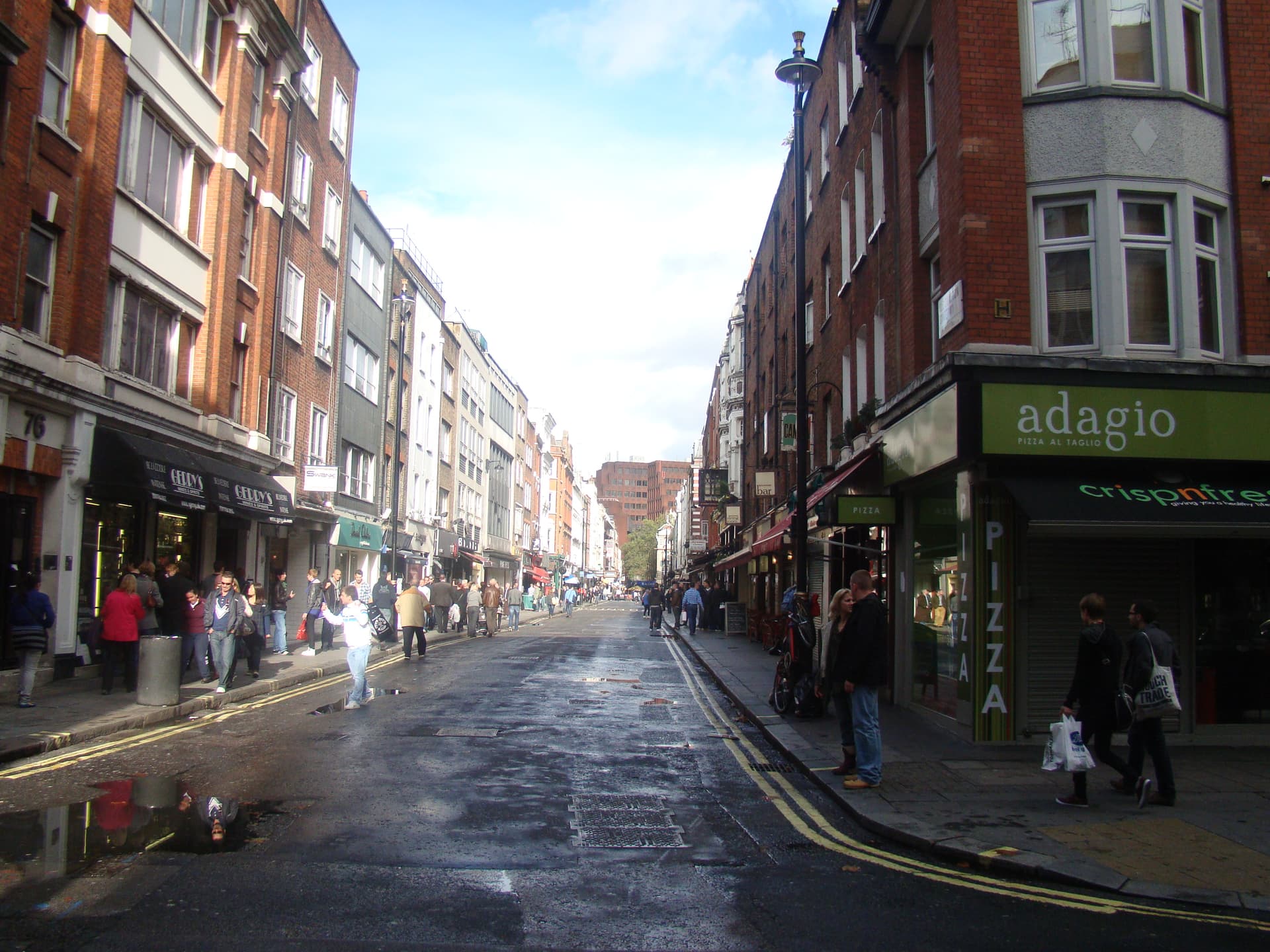 Pedestrians walk along a damp Soho street lined with red-brick buildings, shopfronts, bars and pizza places under blue sky