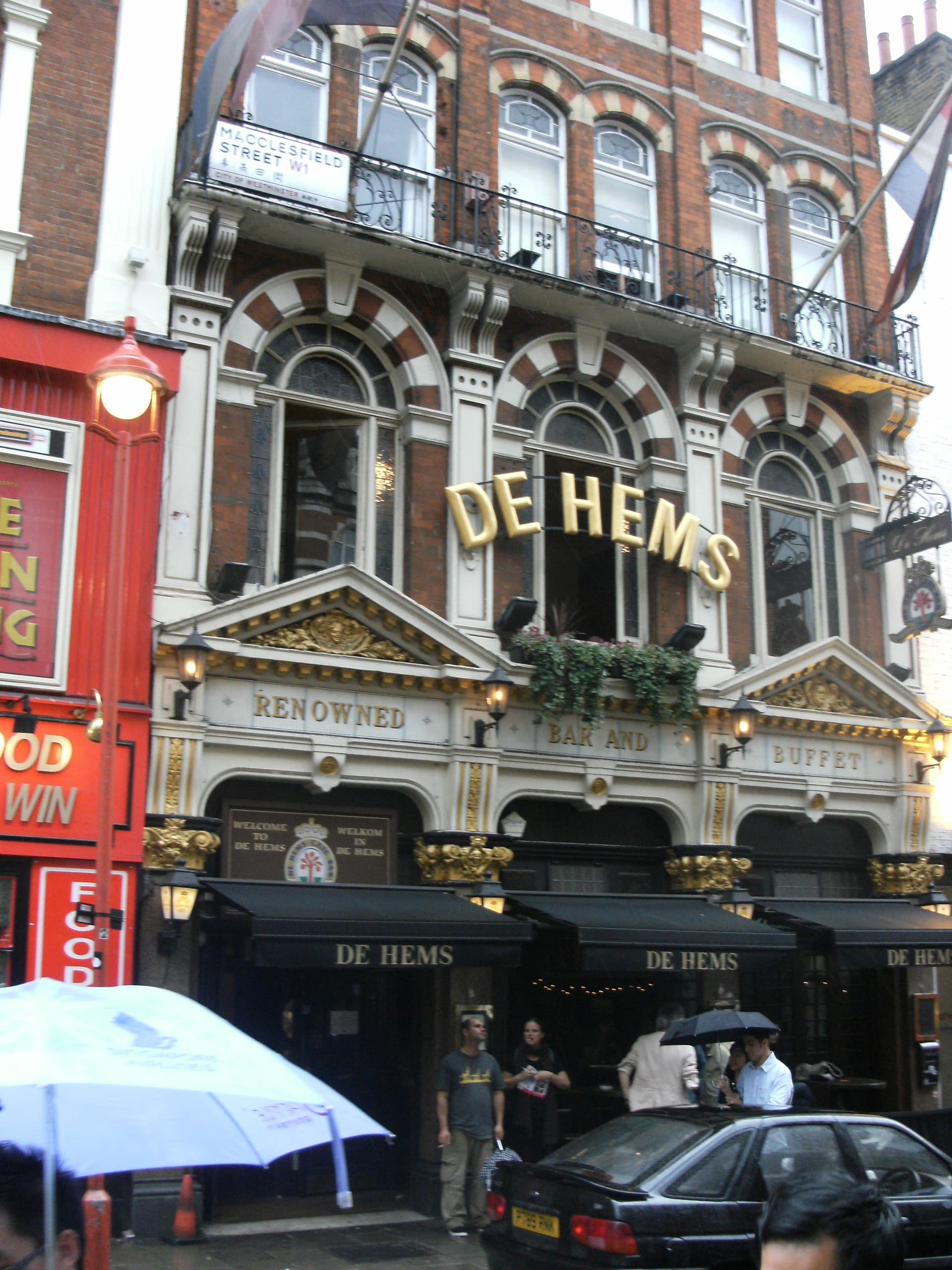 Ornate red-brick and gilded frontage of De Hems pub on Macclesfield Street, Soho, with black awnings on a rainy day