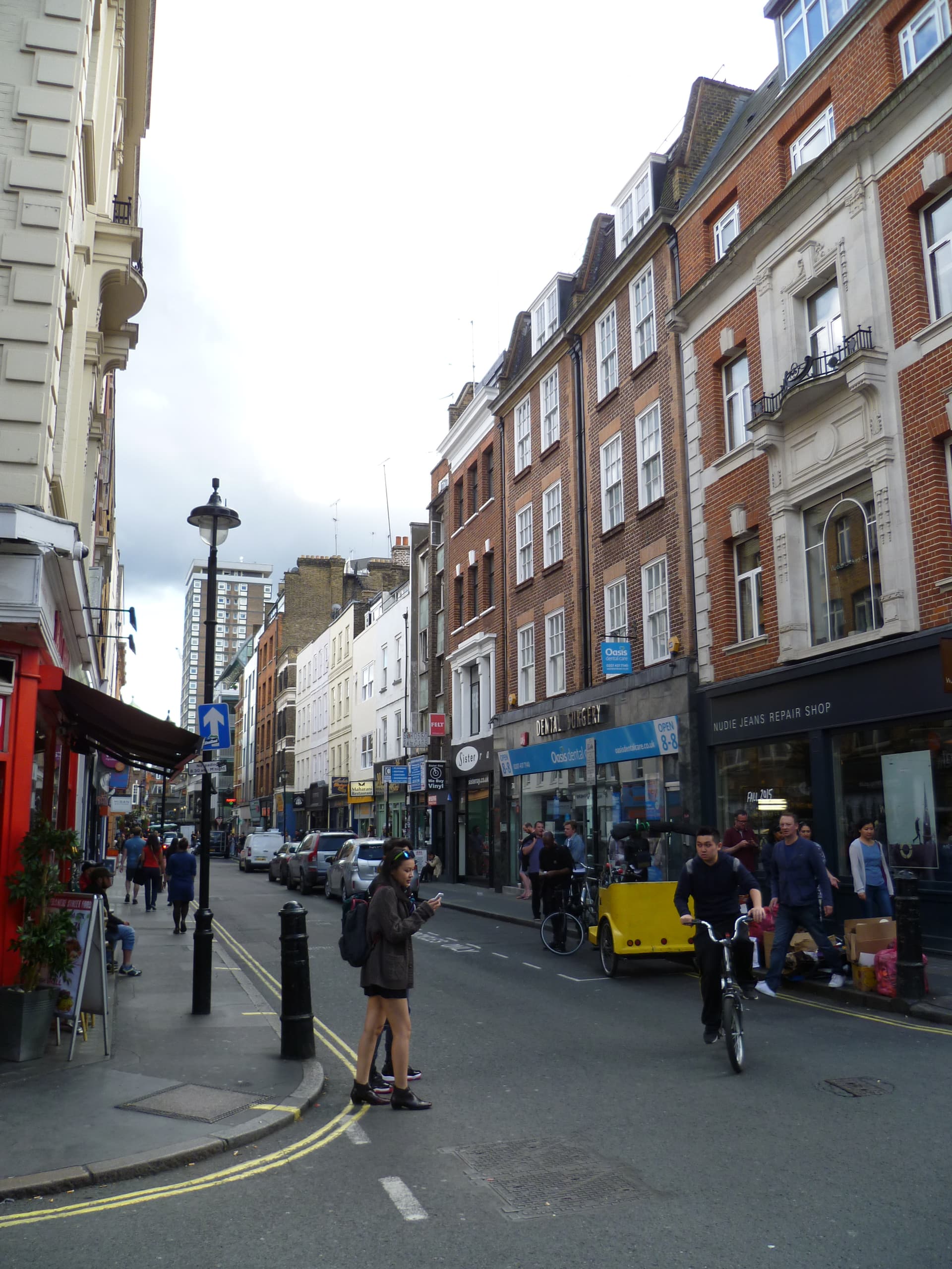 Pedestrians and a cyclist on a Soho street lined with red-brick buildings, shopfronts and parked cars
