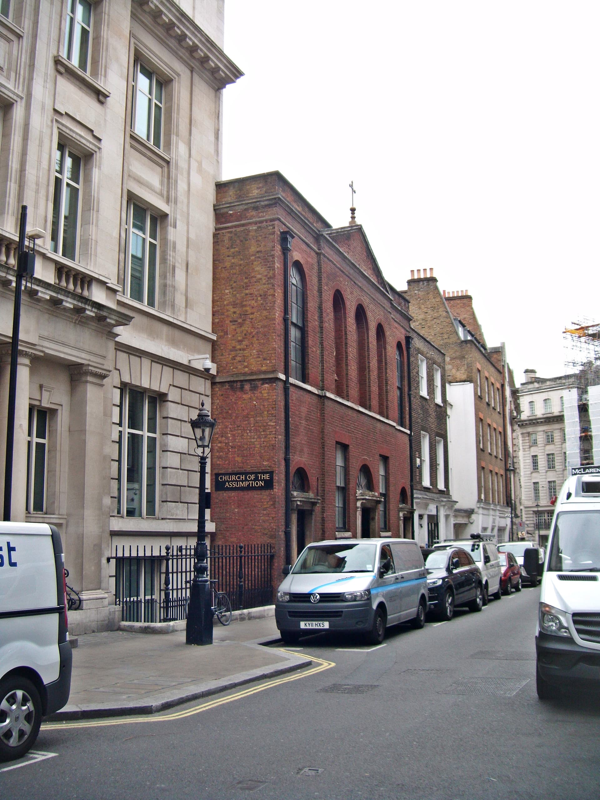 Red brick Church of the Assumption beside stone and brick buildings on a Soho street with parked vans