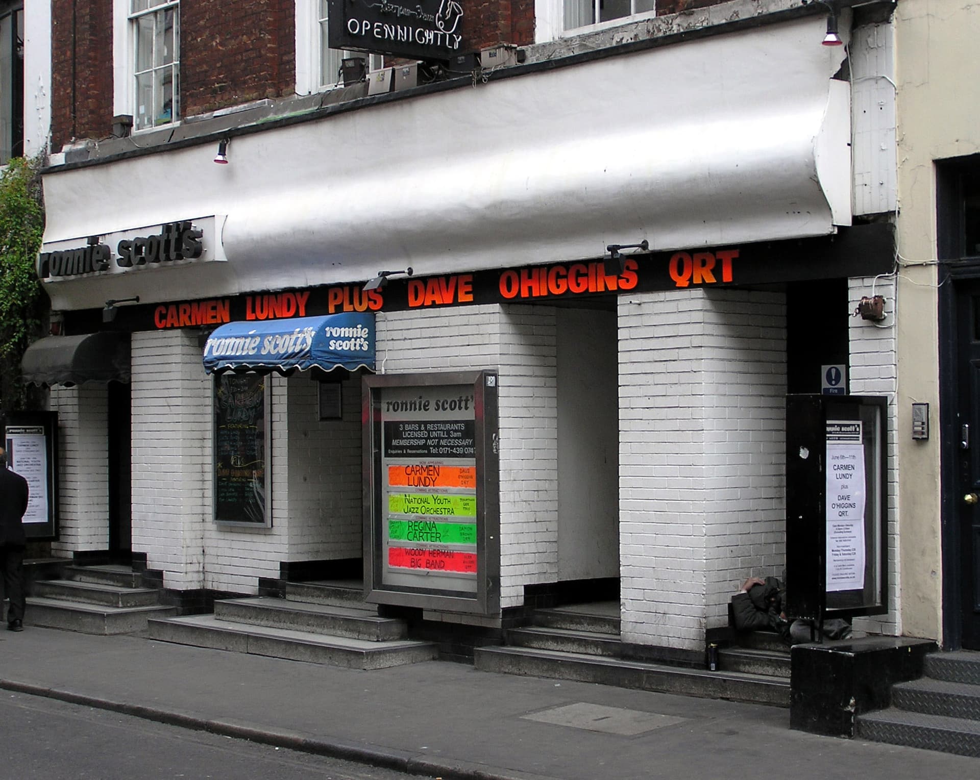 White-tiled frontage of Ronnie Scott's jazz club in Soho, with blue awning, listings boards and orange lettering above the entrances