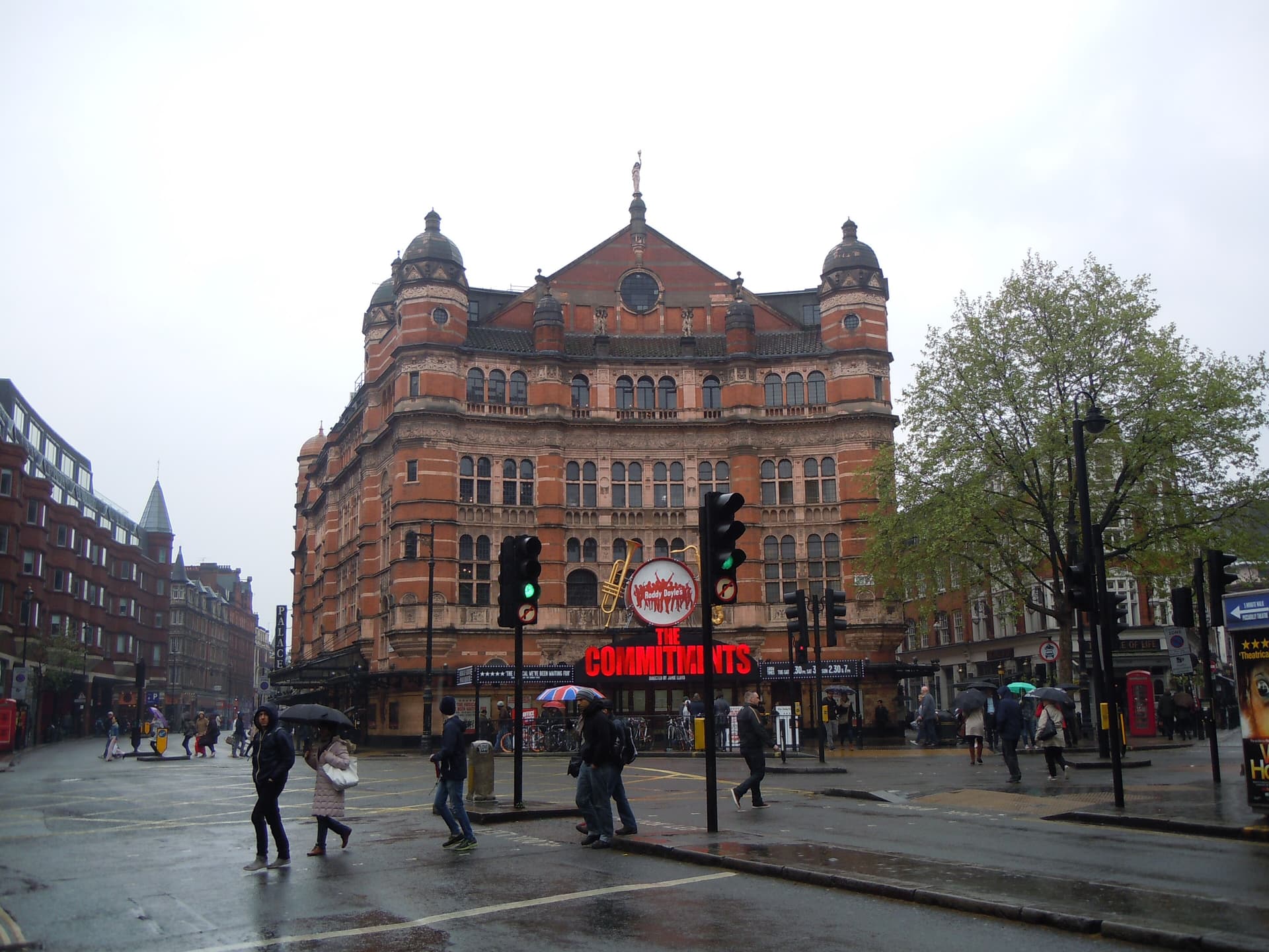 Red-brick Palace Theatre at Cambridge Circus on a wet day, with pedestrians, umbrellas and traffic lights in front