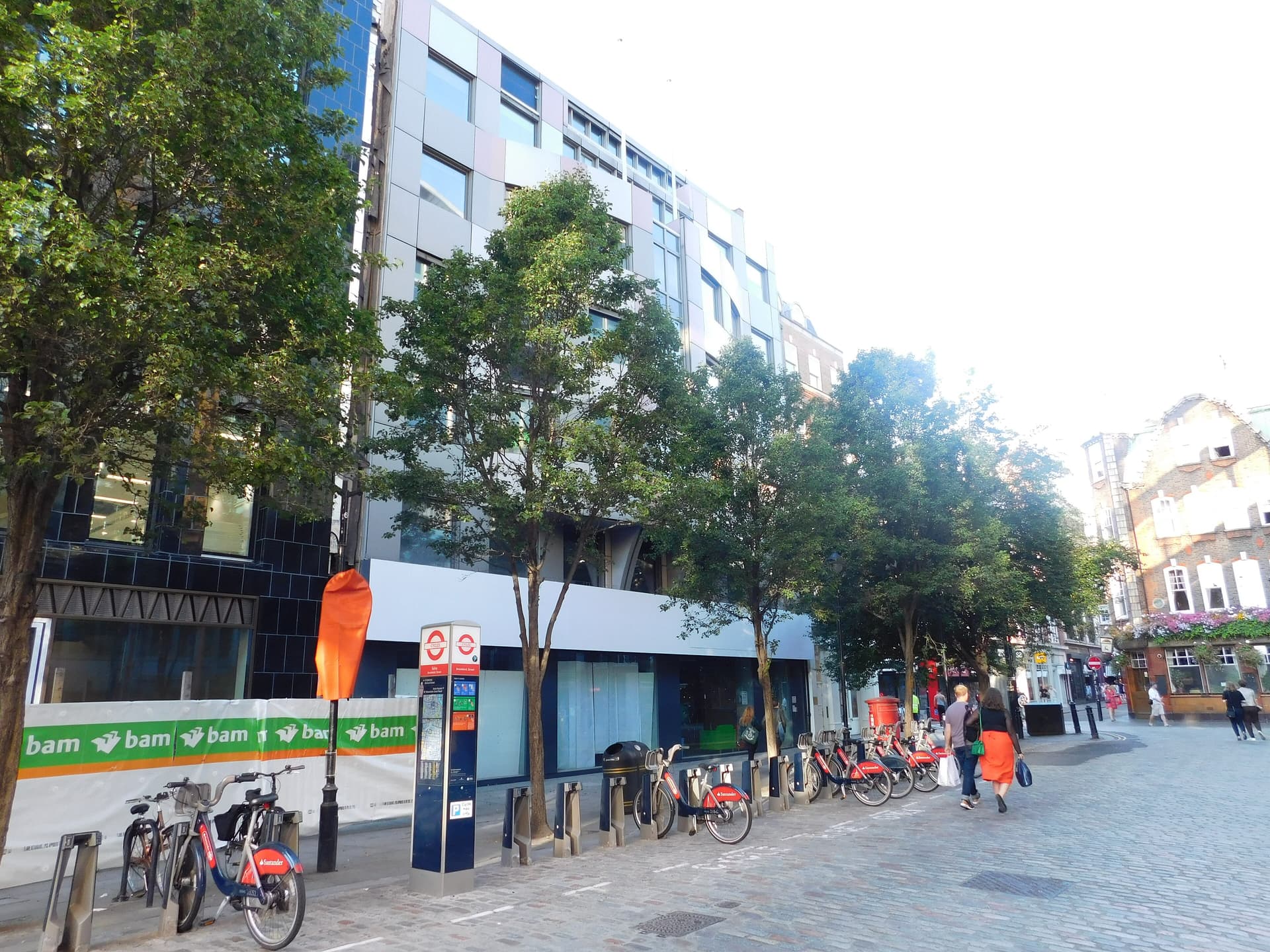 Cobbled Soho street with Santander cycle hire docking station, plane trees, a modern panelled building under hoardings and a pub beyond