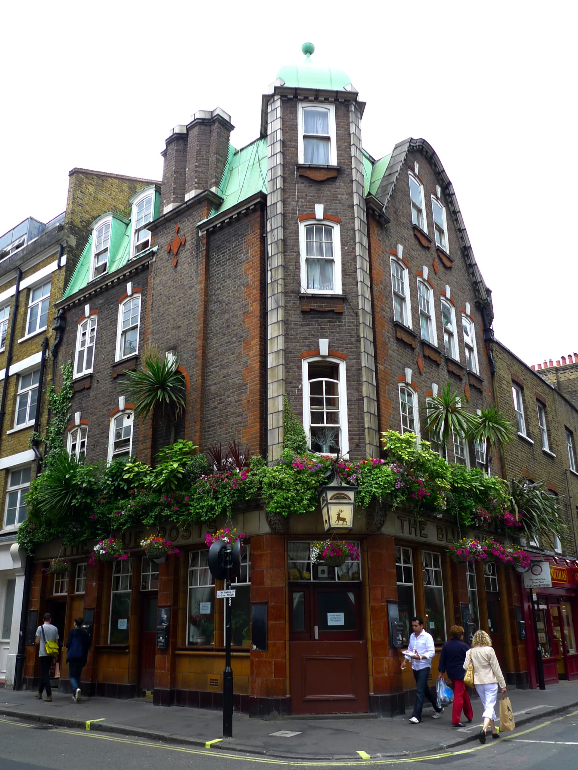 Corner pub with tiled red-brown ground floor, green copper turret and hanging flower baskets on a Soho street