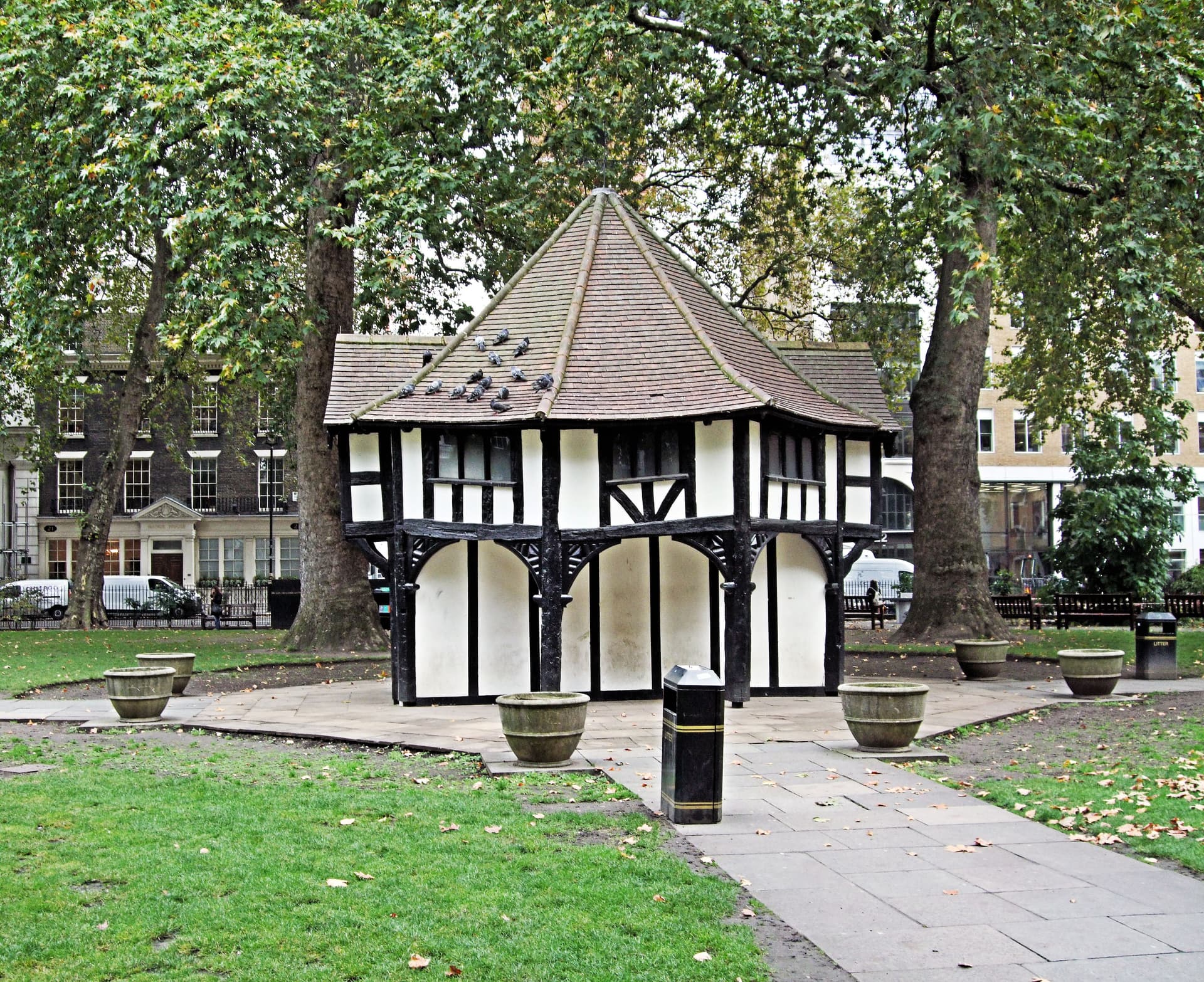 Mock Tudor timber-framed garden keeper's hut with pigeons on its tiled roof, among plane trees in Soho Square