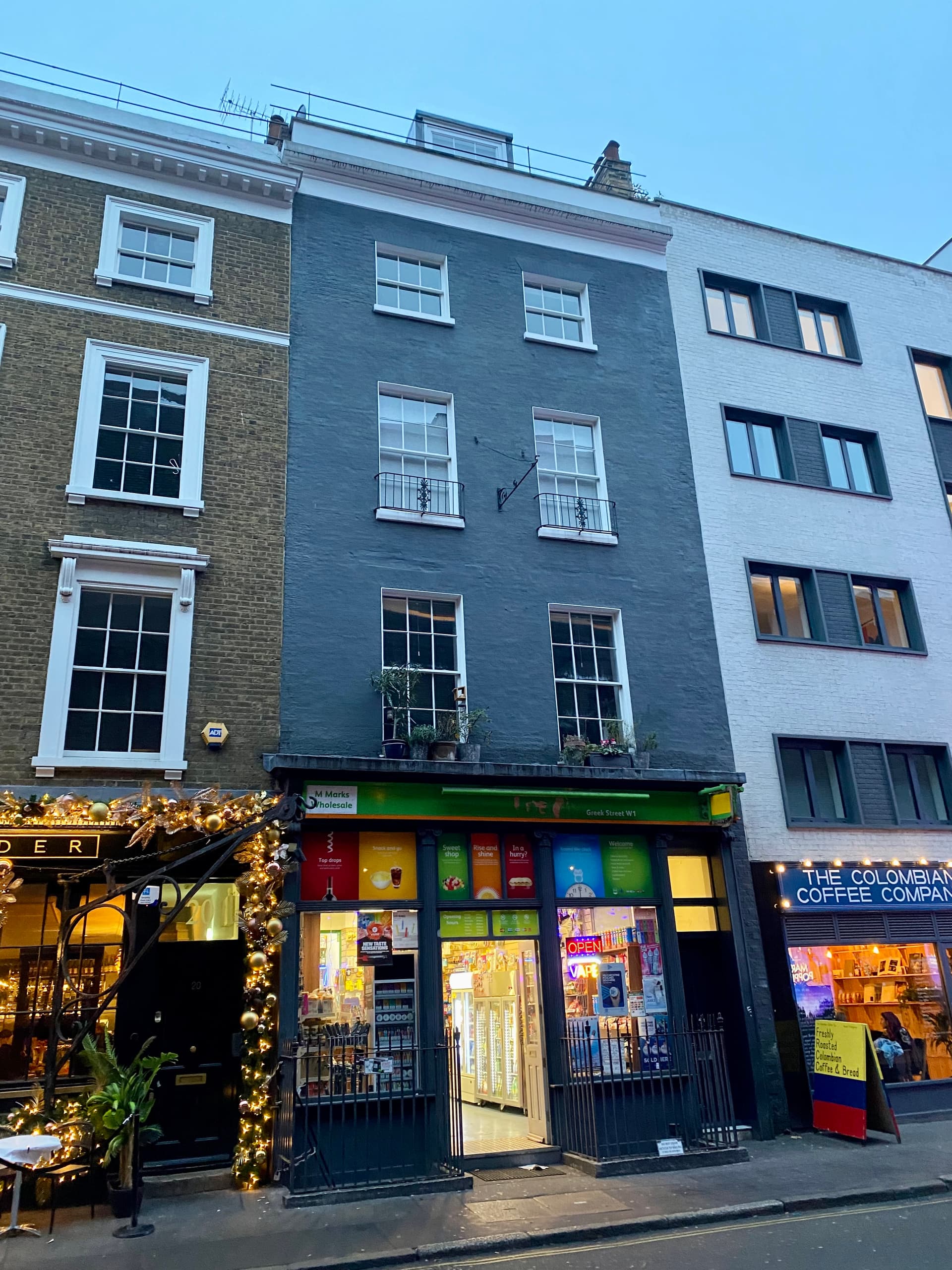 Painted grey Georgian townhouse with a corner-shop front on Greek Street, Soho, between festive-lit and modern buildings