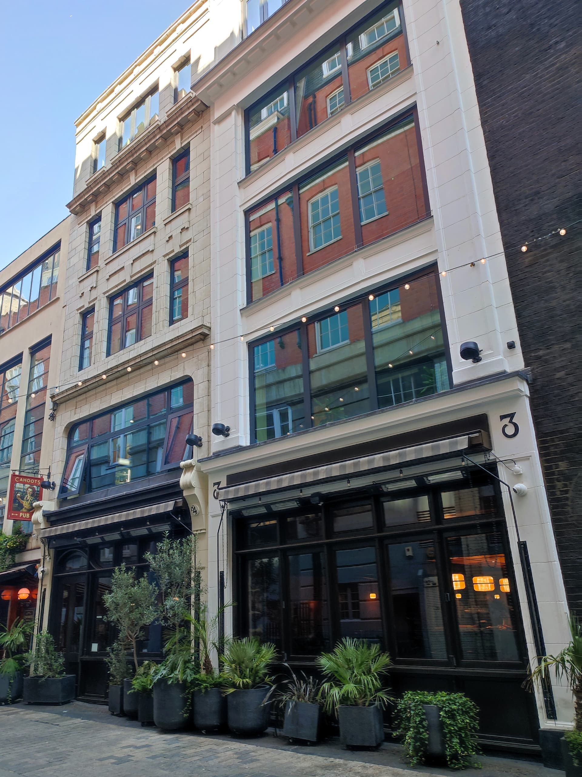 Cream-painted Edwardian buildings with large reflective windows, striped awnings and potted plants along a Soho street, with a Cahoots pub sign