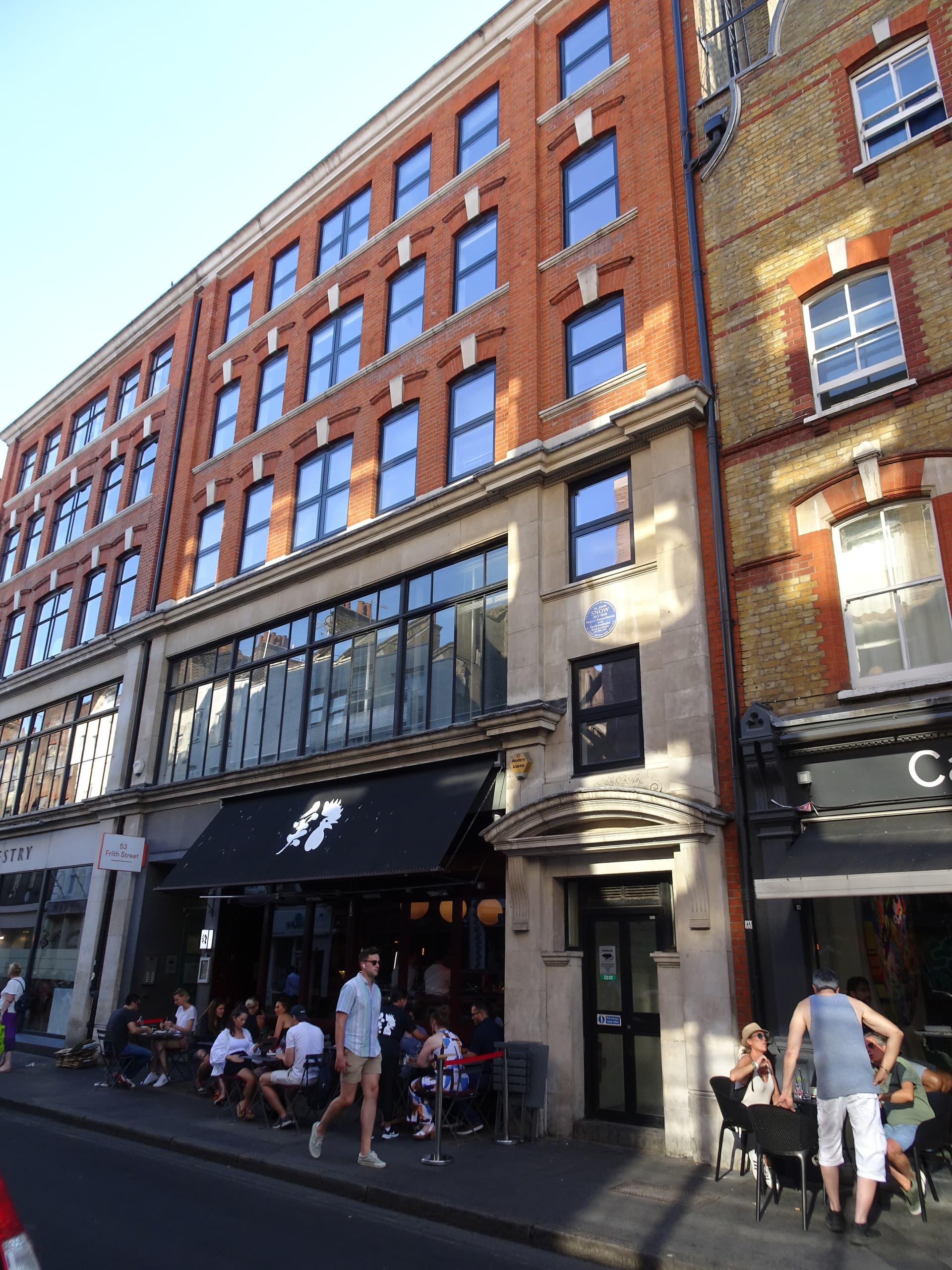 Red-brick Victorian building on Frith Street, Soho, with a blue plaque and diners at pavement tables